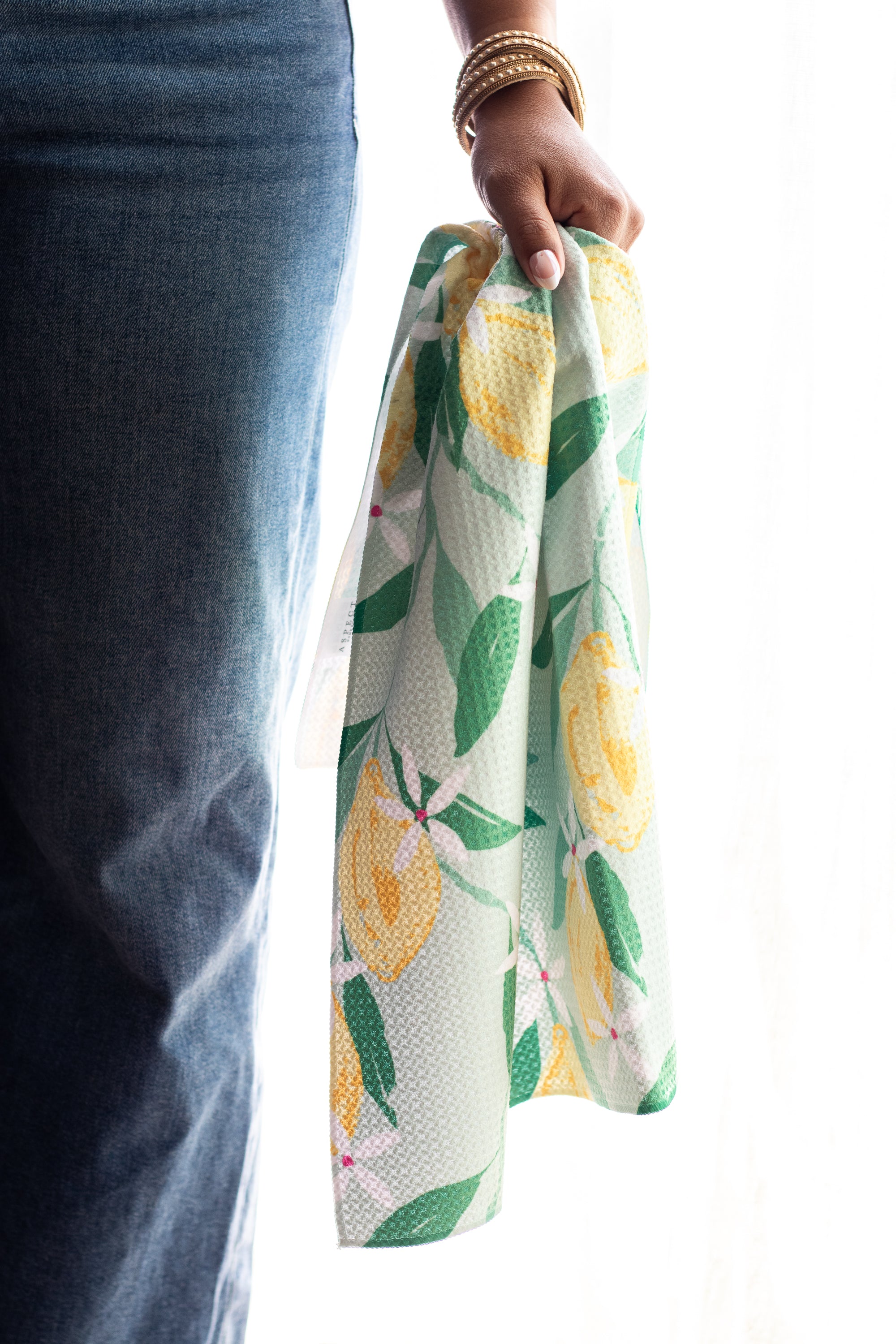 A person wearing blue jeans holds a mint green woven waffle-weave kitchen tea towel with a yellow lemon-and-green leaf pattern against a bright white background.