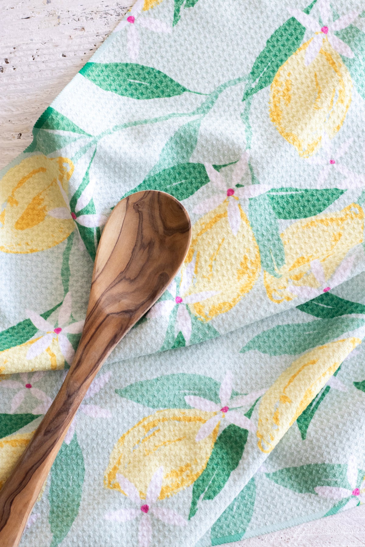 A close-up of a waffle-weave kitchen tea towel with a watercolor lemon print, featuring yellow lemons, green leaves, and small white-and-pink flowers on a mint green background. A wooden spoon is placed diagonally across the towel.