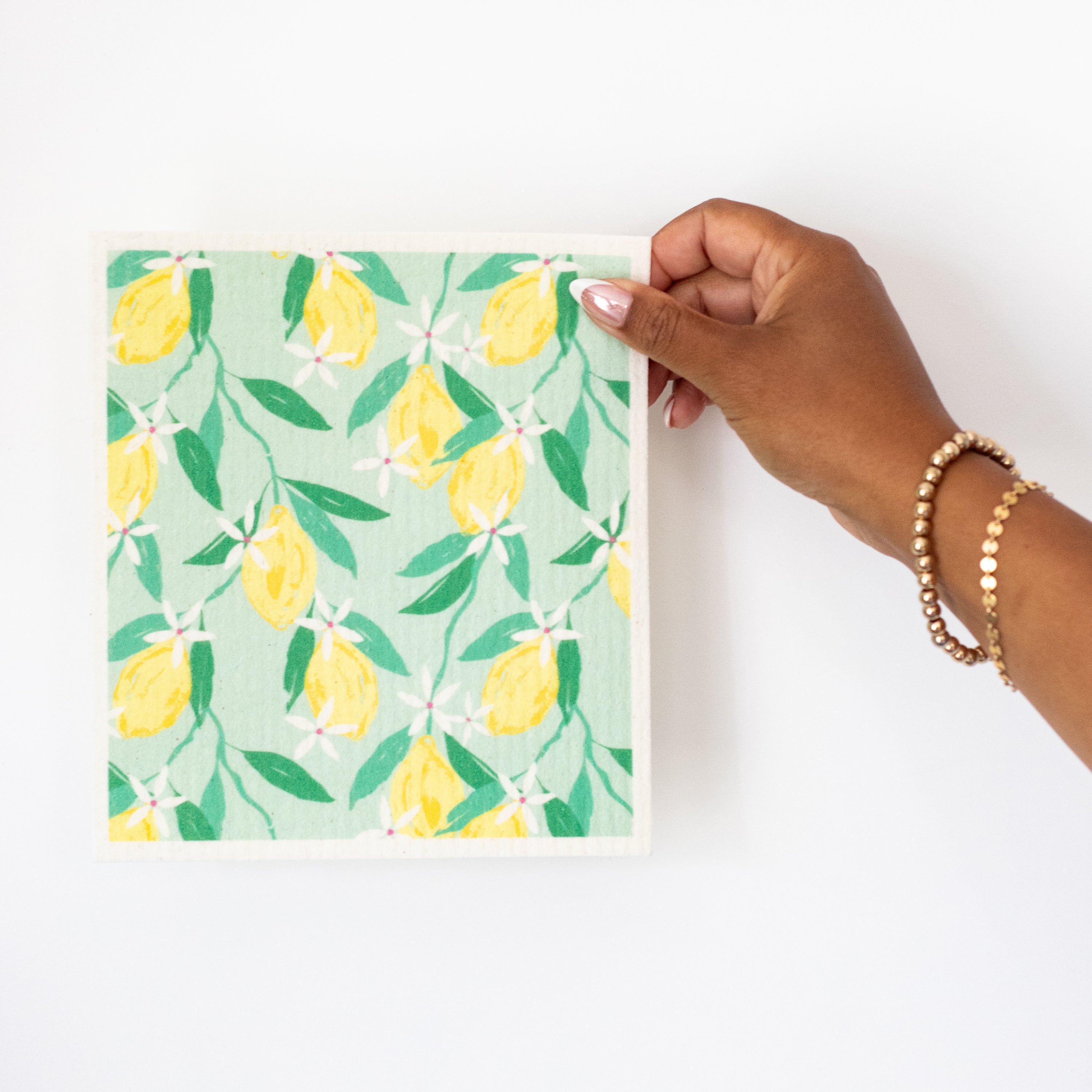 A hand holding a Swedish dishcloth with a yellow lemon and green leaf pattern on a mint-green background, set against a plain white wall.