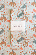 A folded waffle-weave kitchen tea towel with a western-themed print of cowboy boots and hats, cowboys riding bucking broncos, kicking bulls, stars, lassos and horseshoes, featuring a white paper belly band label in the center that reads 'ASPECT HOME GOODS', laid on a background of the same pattern.