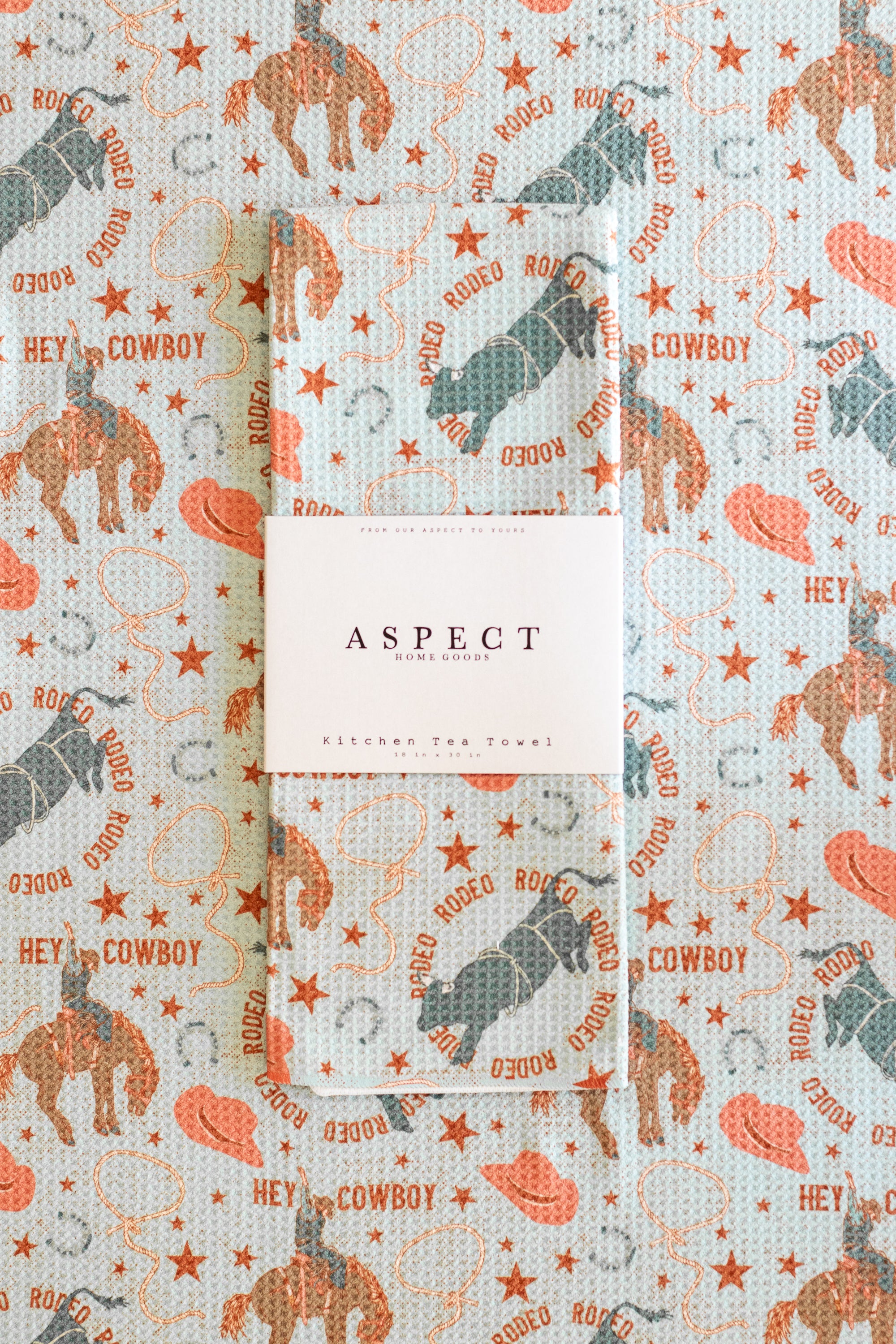 A folded waffle-weave kitchen tea towel with a western-themed print of cowboy boots and hats, cowboys riding bucking broncos, kicking bulls, stars, lassos and horseshoes, featuring a white paper belly band label in the center that reads 'ASPECT HOME GOODS', laid on a background of the same pattern.