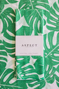 A folded kitchen tea towel with a green and white monstera leaf pattern, wrapped with a white paper belly band labeled 'ASPECT HOME GOODS' and 'KITCHEN TEA TOWEL'.