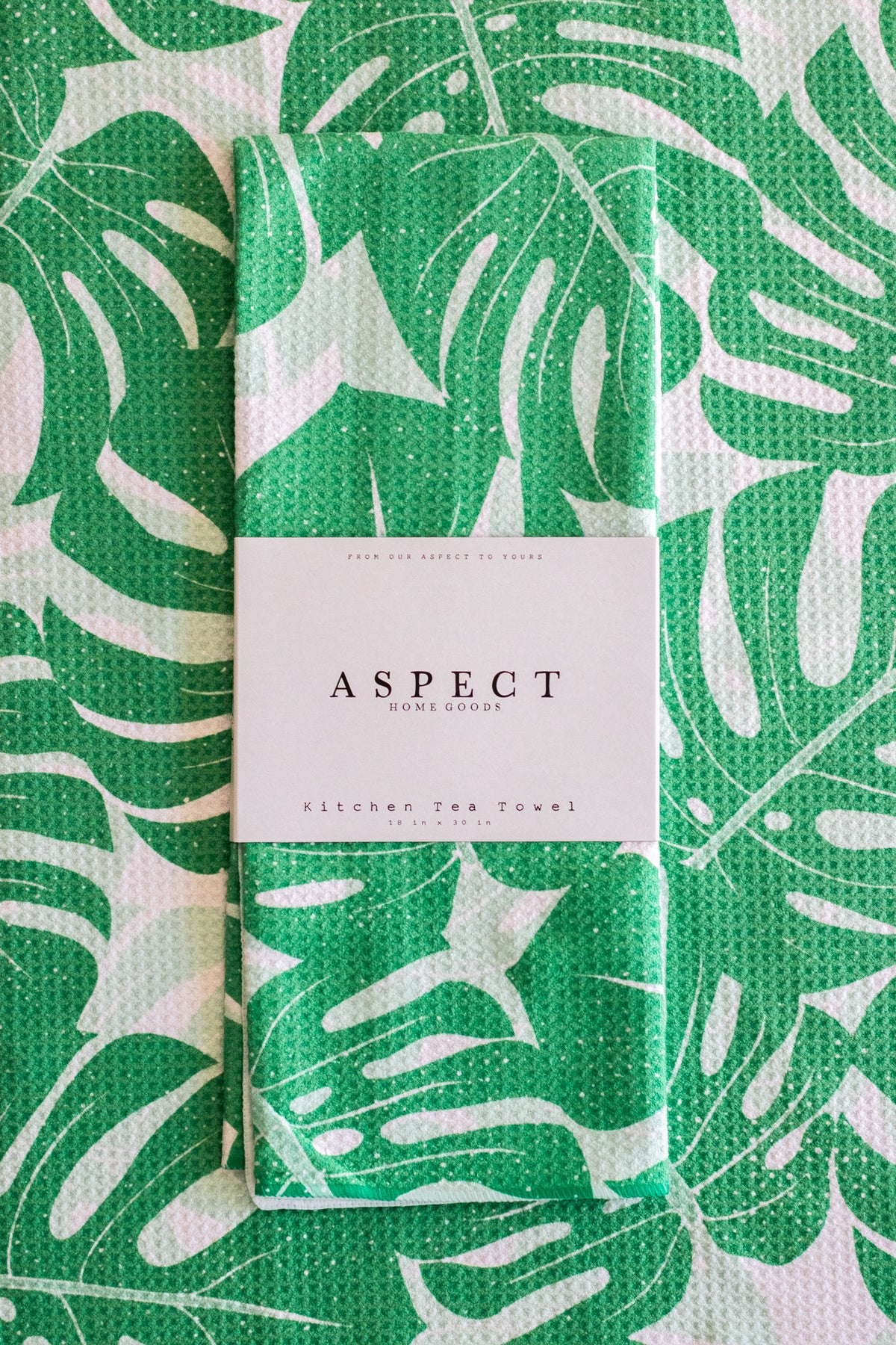 A folded kitchen tea towel with a green and white monstera leaf pattern, wrapped with a white paper belly band labeled 'ASPECT HOME GOODS' and 'KITCHEN TEA TOWEL'.