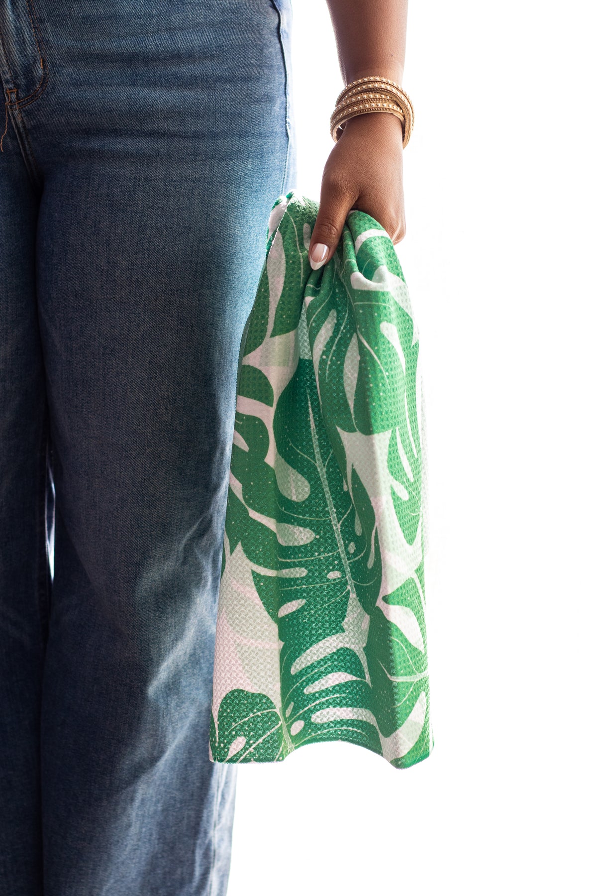 A person wearing blue jeans holds a green-and-white tropical monstera leaf patterned towel against a white background.