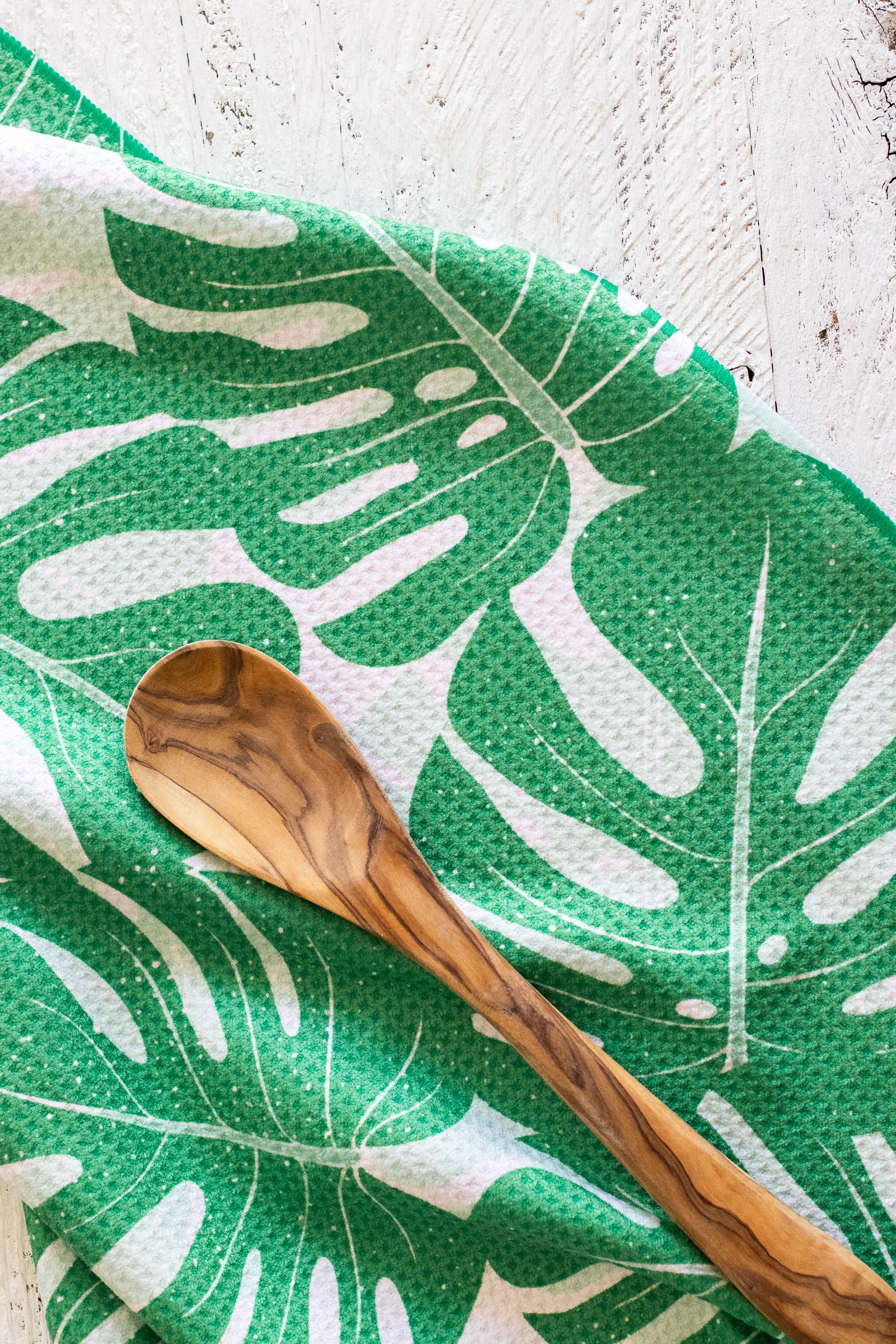 A green-and-white tropical monstera leaf patterned kitchen tea towel with a wooden spoon resting on it, set against a white rustic wooden background.