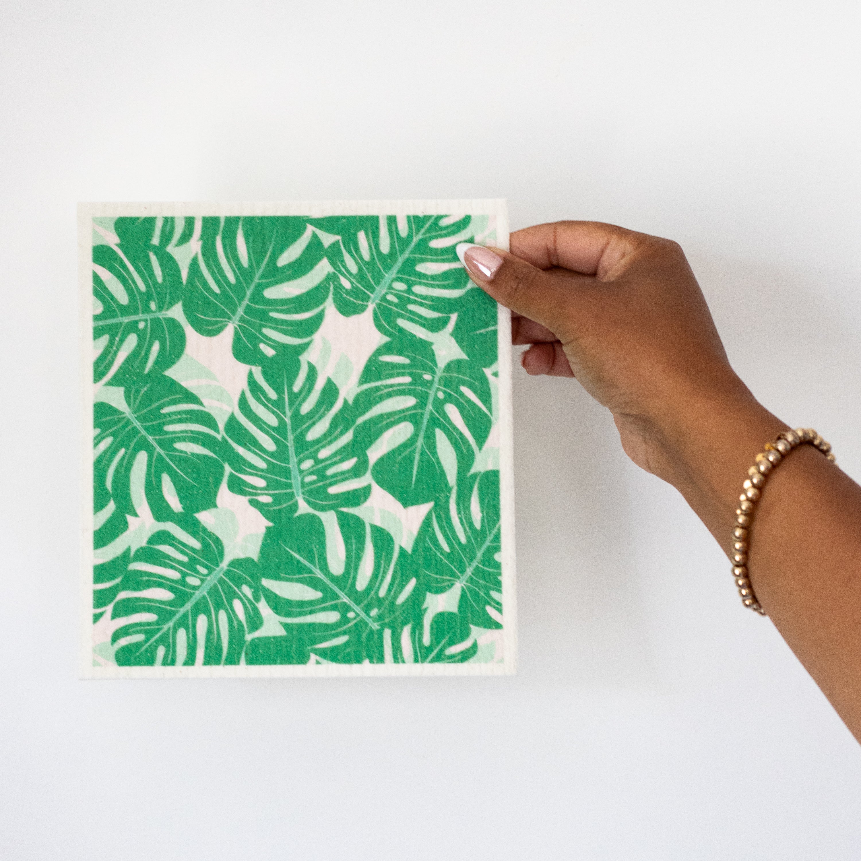 A hand holding a Swedish dishcloth featuring a green tropical monstera leaf pattern against a plain white background.