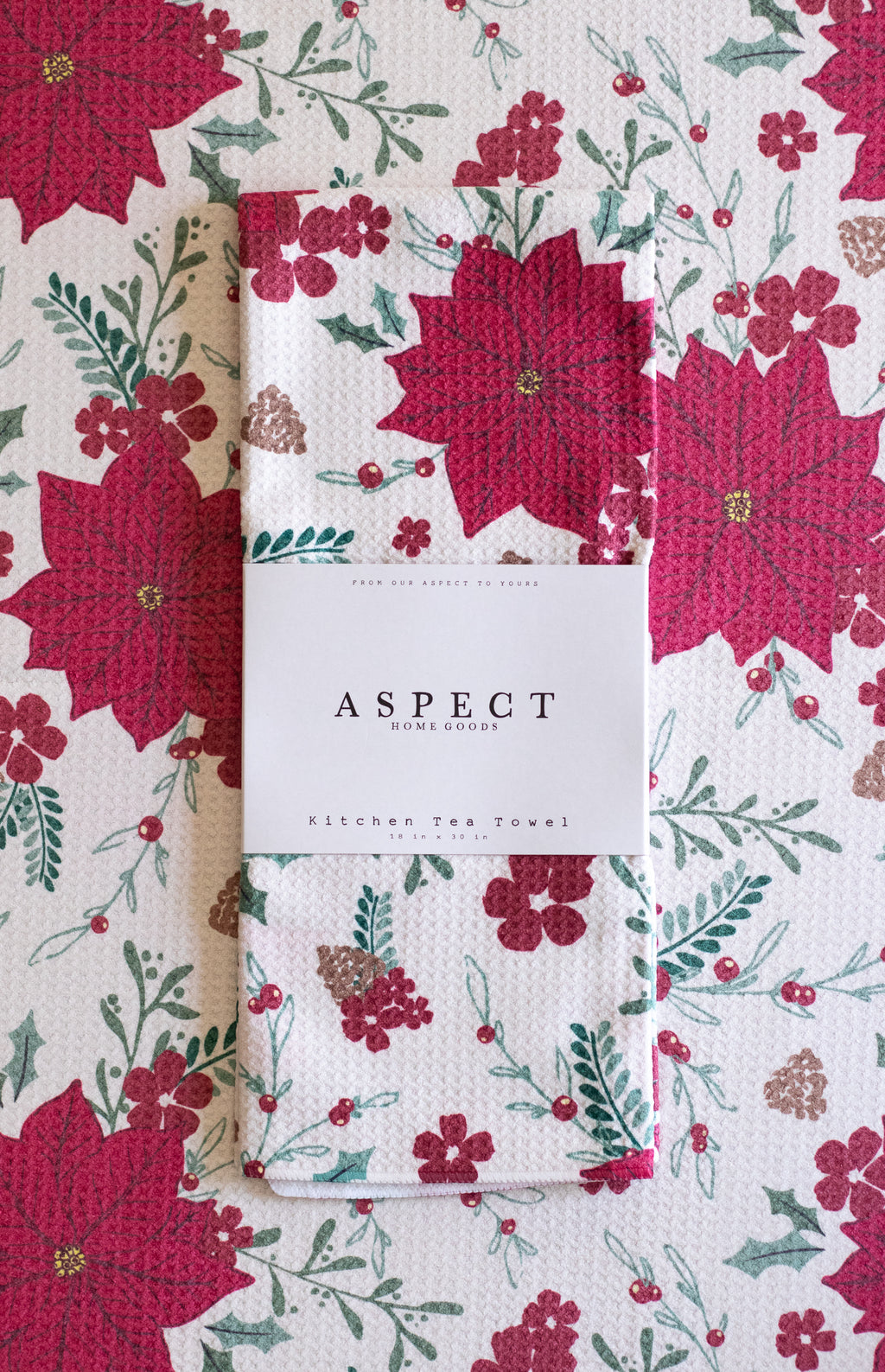 A folded waffle-weave kitchen tea towel with a festive red, green, and white poinsettia, berry, and pinecone pattern, wrapped in a white paper belly band labeled 'ASPECT HOME GOODS'. The towel is on top of another towel with the same pattern.