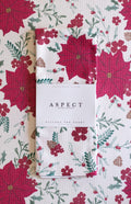 A folded waffle-weave kitchen tea towel with a festive red, green, and white poinsettia, berry, and pinecone pattern, wrapped in a white paper belly band labeled 'ASPECT HOME GOODS'. The towel is on top of another towel with the same pattern.