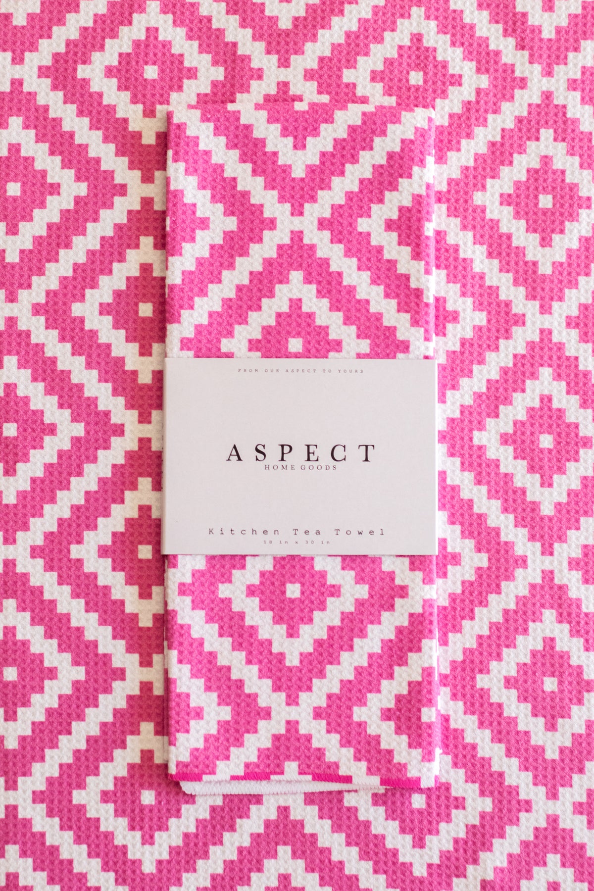 A folded kitchen tea towel with a pink and white geometric diamond pattern, featuring a white paper belly band that reads 'ASPECT HOME GOODS' and 'Kitchen Tea Towel'. The background is a matching flat-lay of the same tea towel pattern.