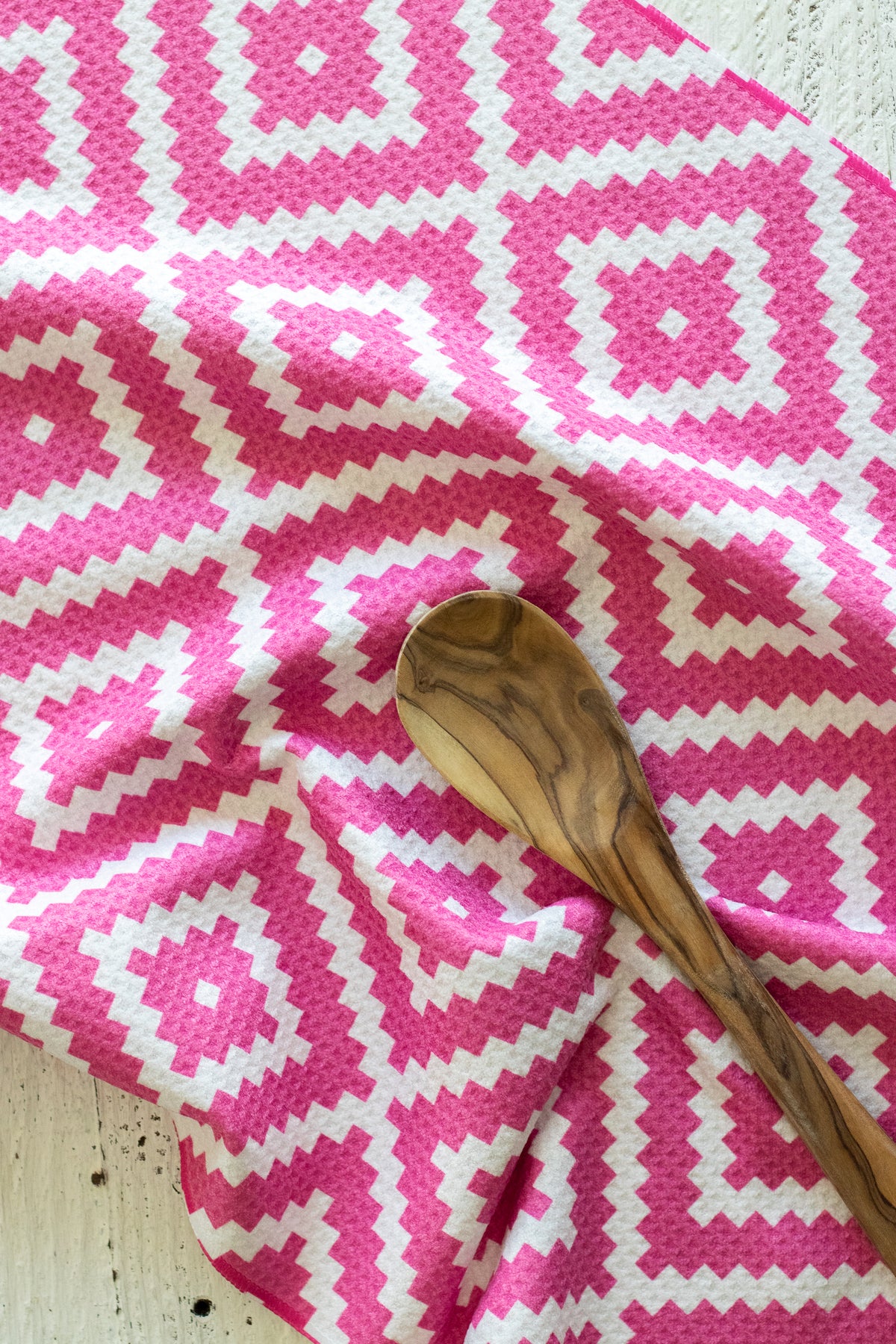 A close-up of a pink and white geometric patterned waffle-weave kitchen tea towel with a wooden spoon resting on it.
