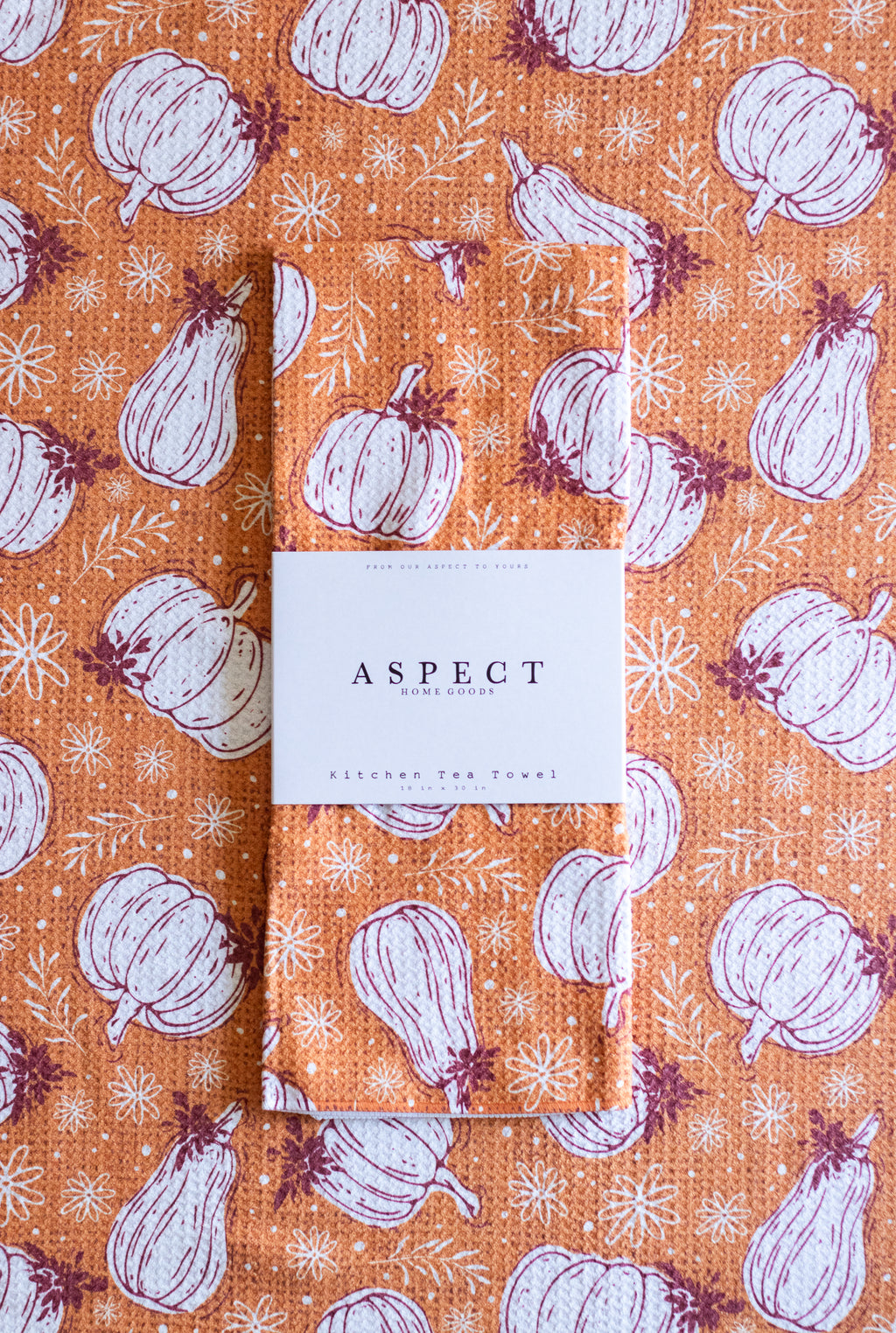 A waffle-weave orange kitchen tea towel with a white and burgundy pumpkin and gourd pattern with flowers, folded with a white paper belly band label that reads 'ASPECT Home Goods Kitchen Tea Towel', laying on a towel with the same pattern.