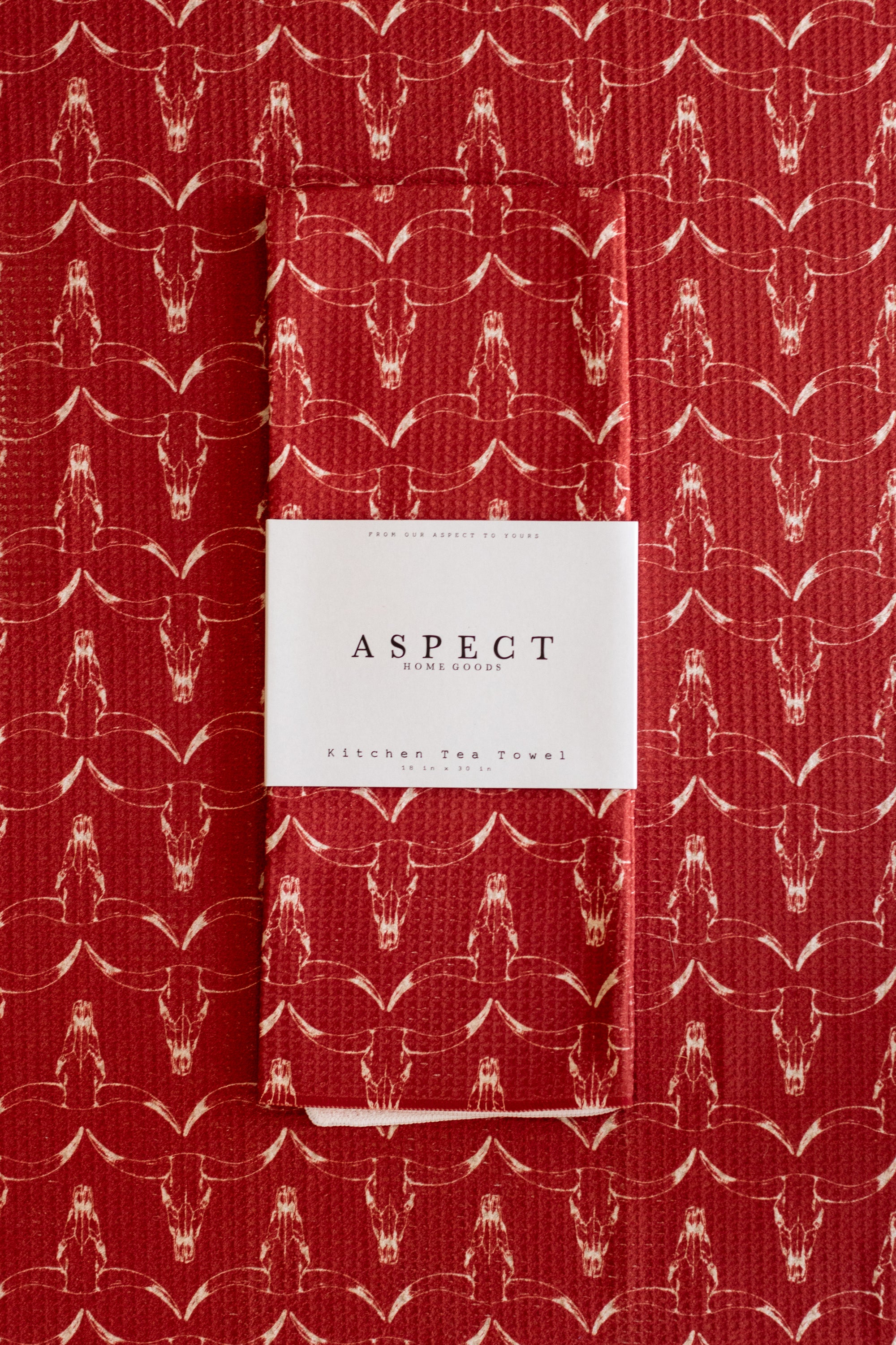 A folded red waffle-weave kitchen tea towel with a repeating hand-drawn white longhorn skull pattern, featuring a white paper belly band label in the center that reads 'ASPECT HOME GOODS' and 'Kitchen Tea Towel', laid on a matching background.