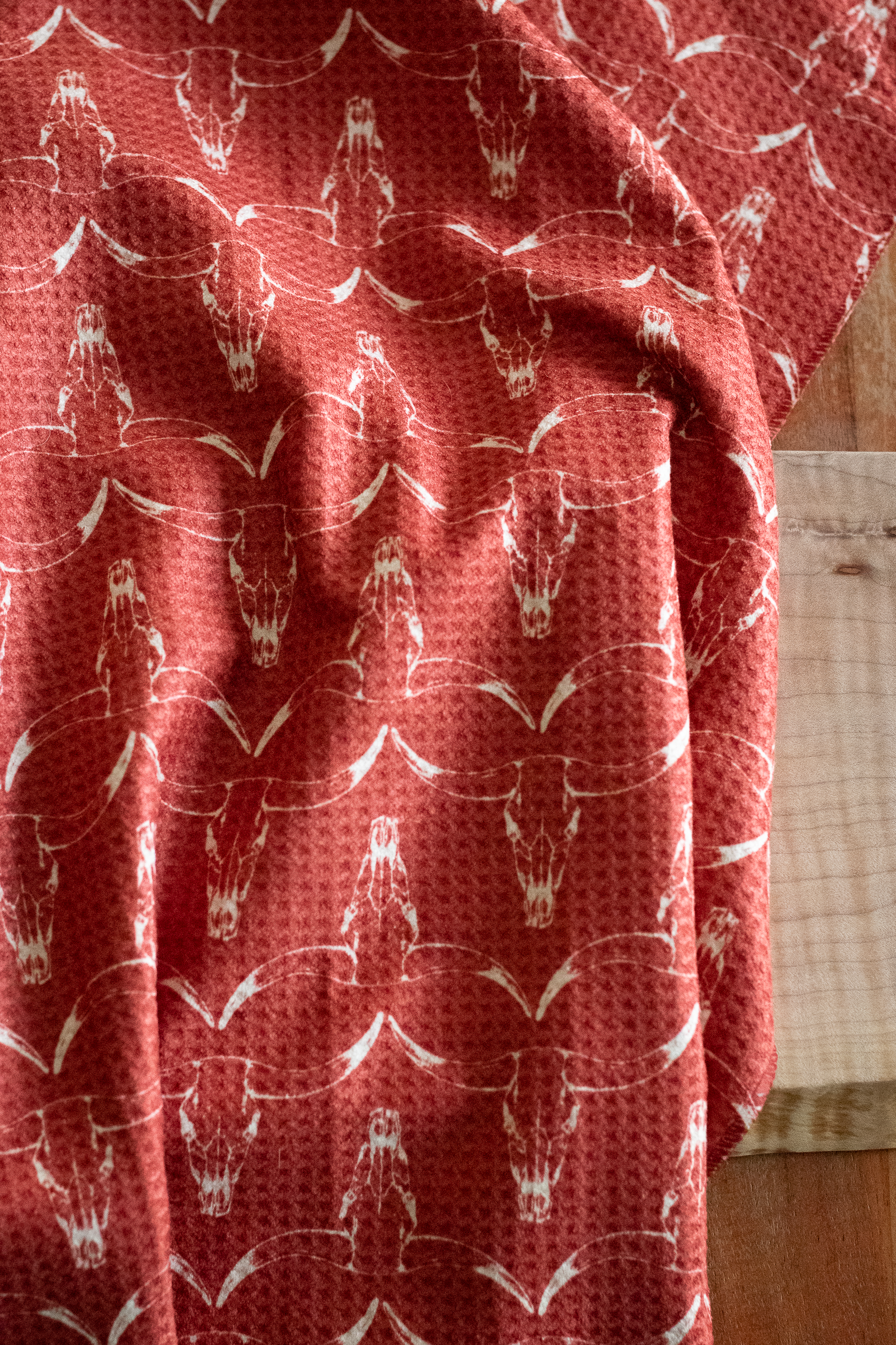 A red waffle-weave kitchen tea towel with a white hand-drawn longhorn cow skull pattern on a wooden surface.