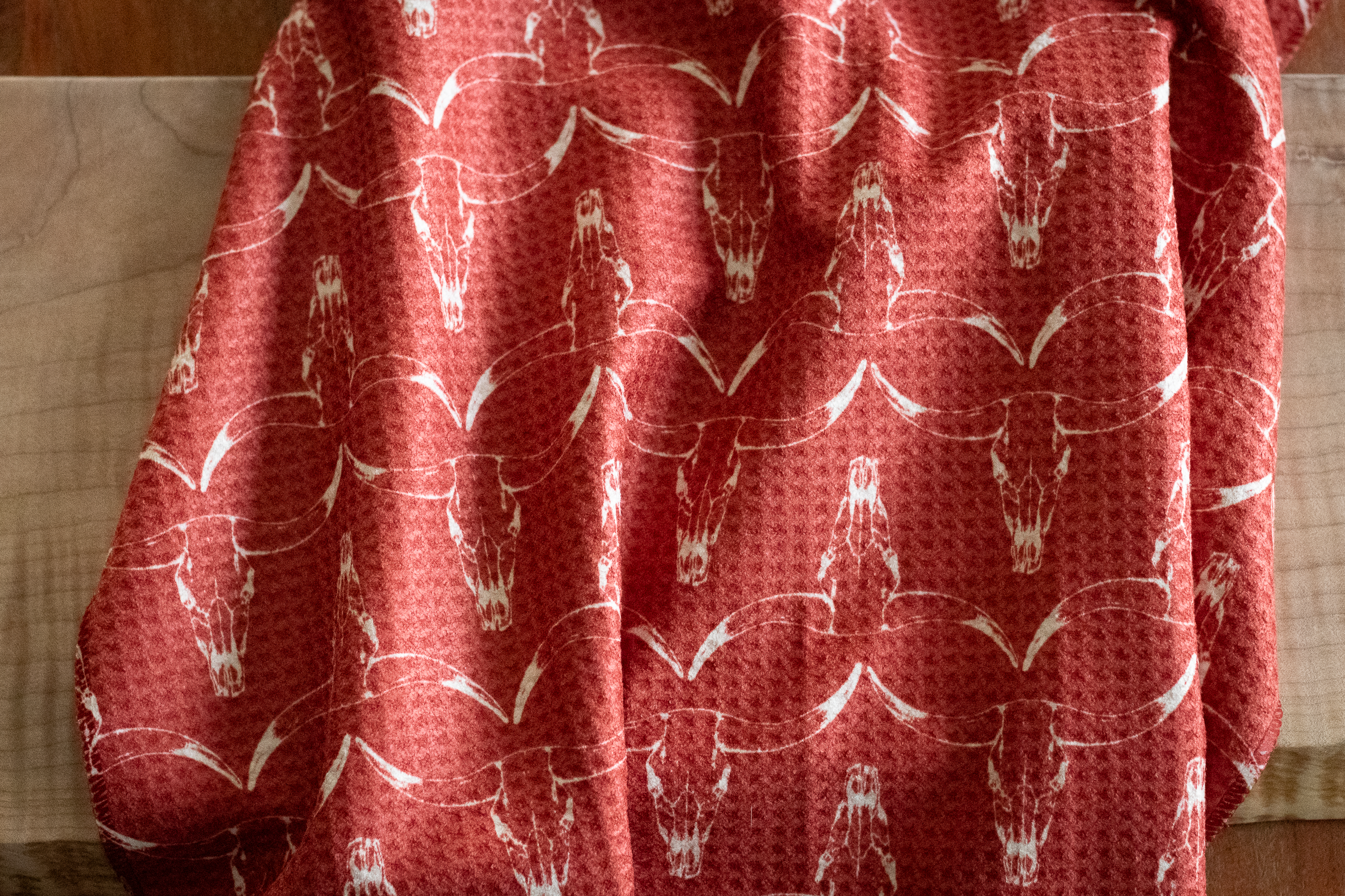 A red waffle-weave kitchen tea towel with a white hand-drawn longhorn cow skull pattern on a wooden surface.