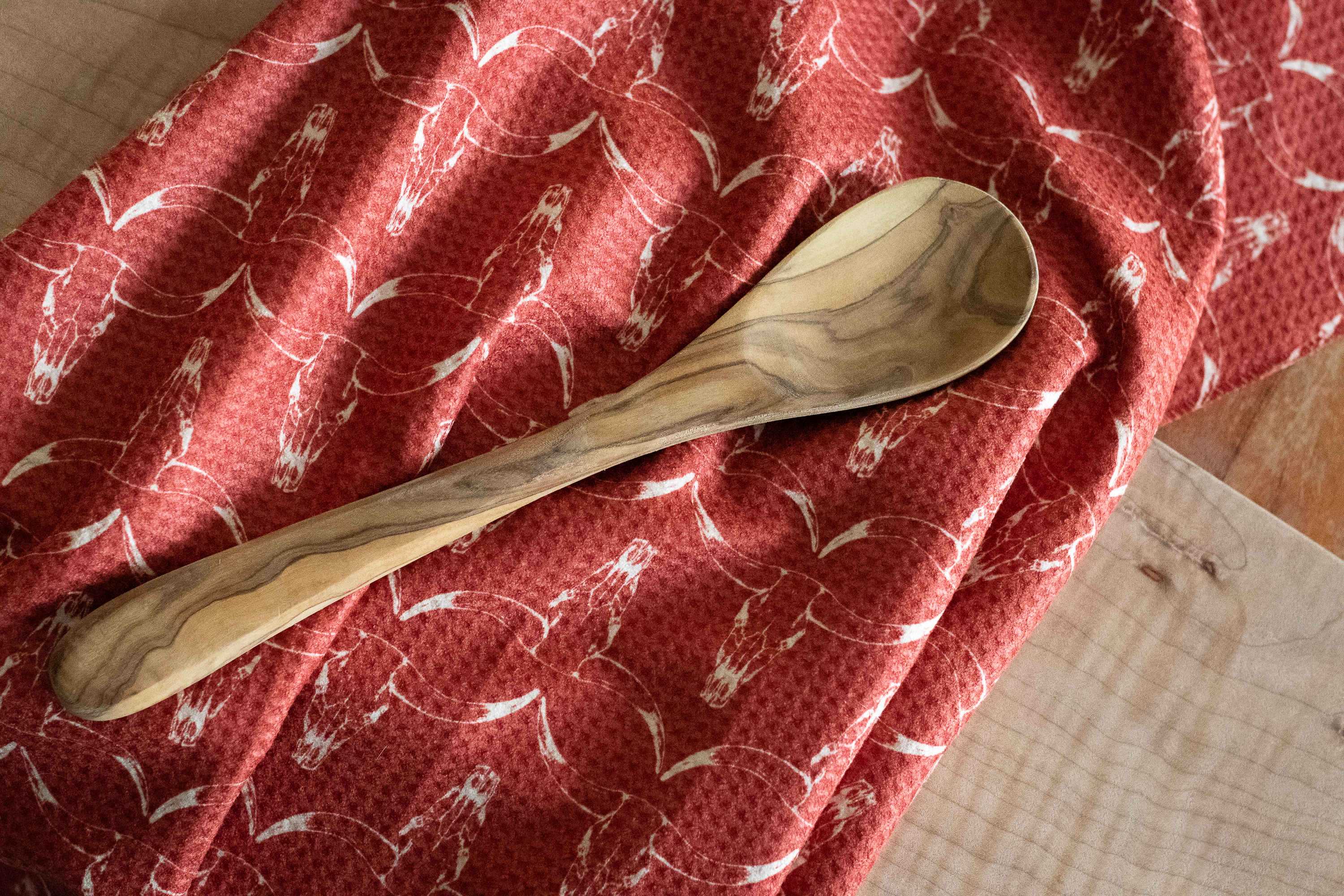 A wooden spoon lying on a red waffle-weave kitchen tea towel with a white hand-drawn longhorn cow skull pattern, on top of a wooden surface.