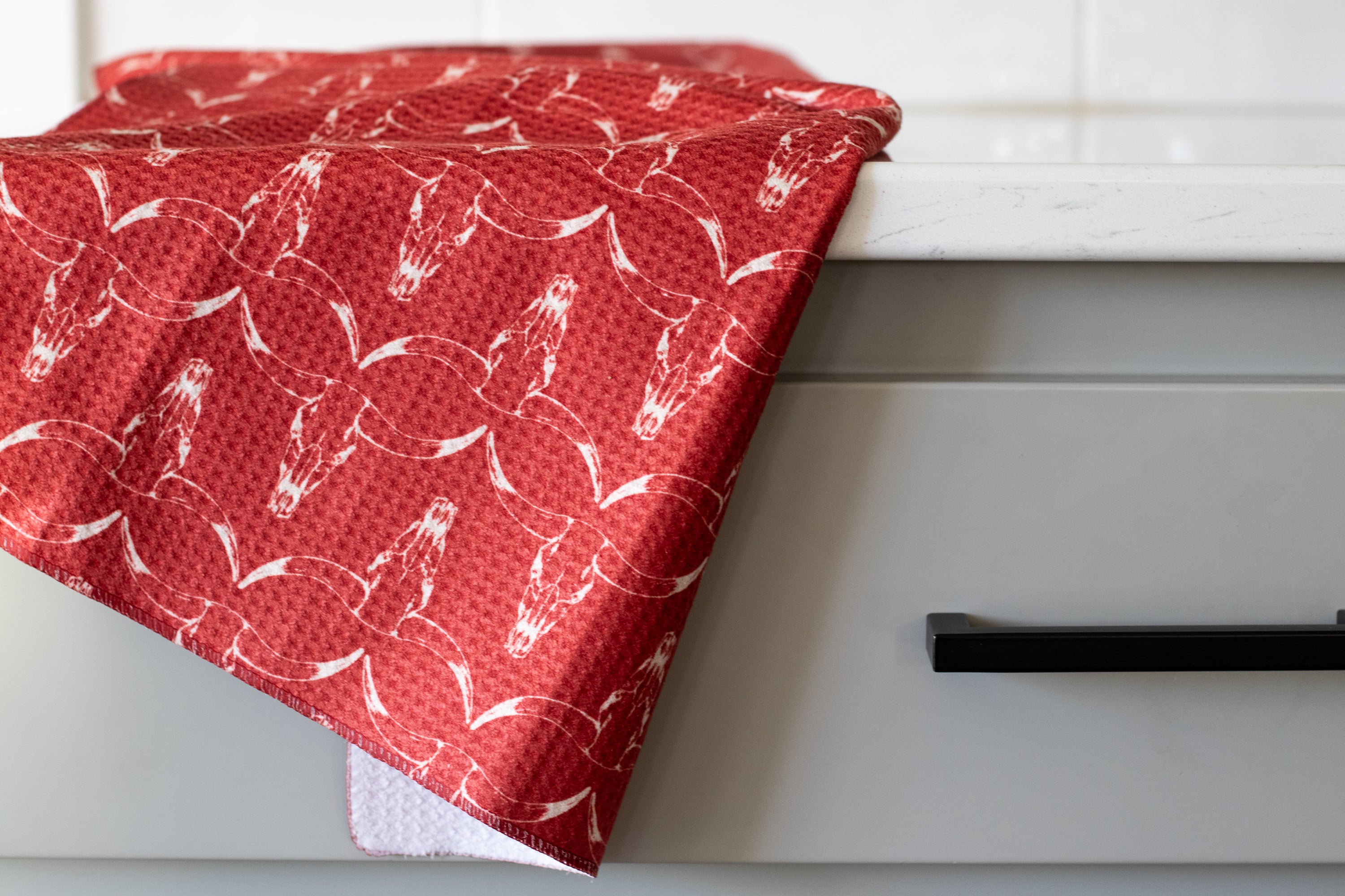 A red waffle-weave kitchen tea towel with a white, hand-drawn longhorn cow skull pattern, draped on a counter in front of a gray drawer with a black handle.