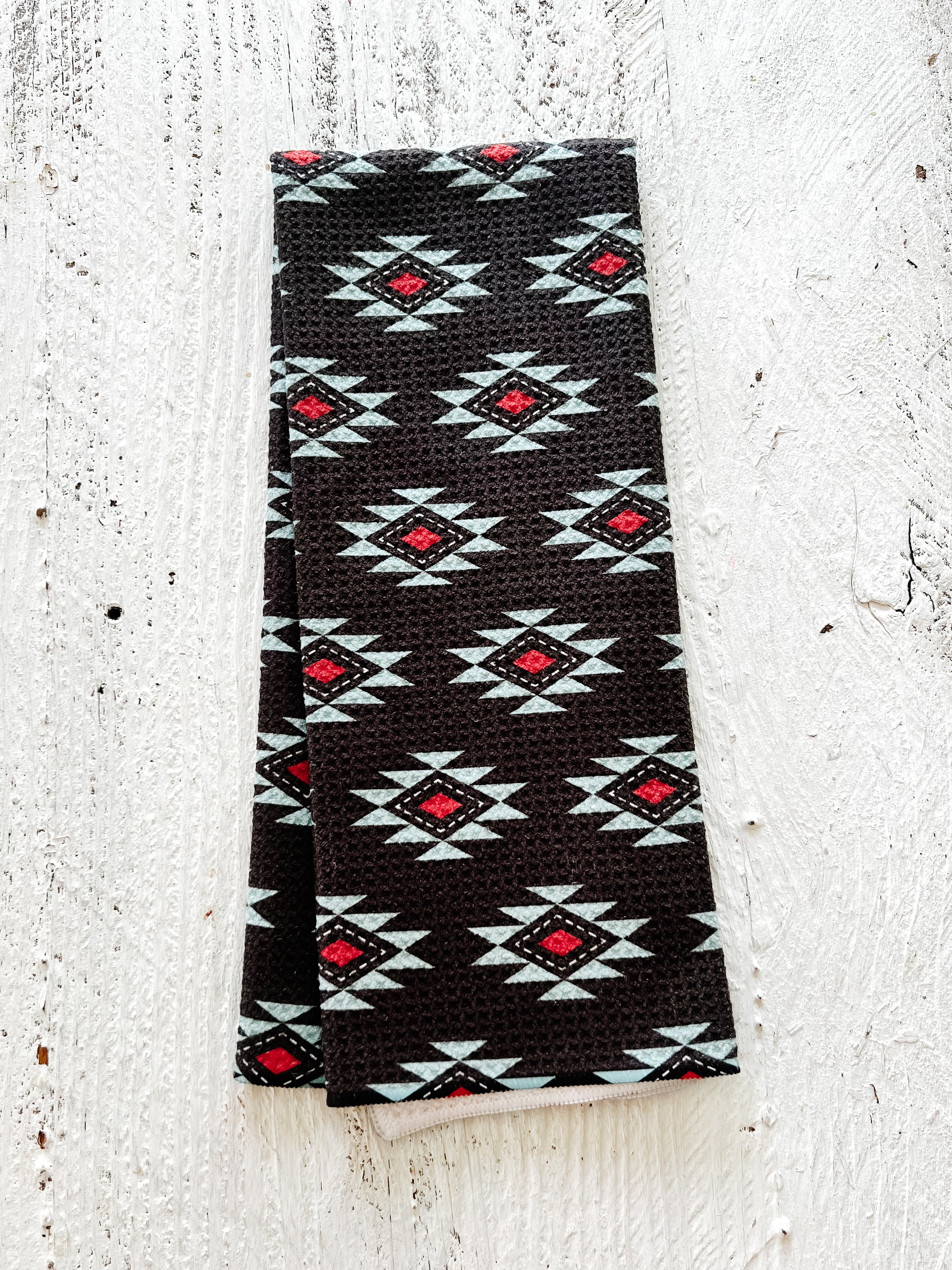 A waffle-weave kitchen tea towel with a Southwestern geometric pattern in blue, red, and white and a black background, folding lengthwise in half, lying on a textured white background.