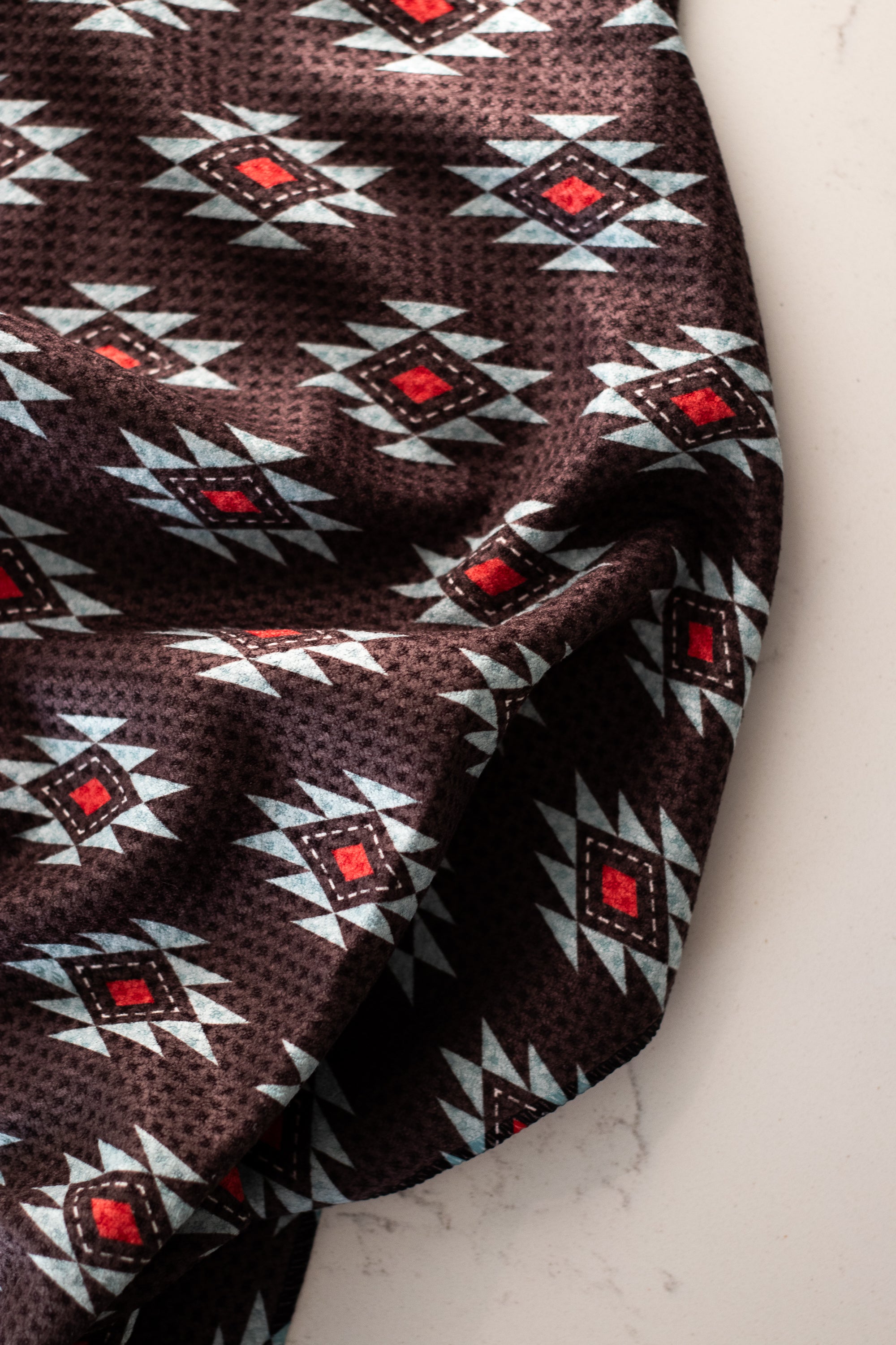 A waffle-weave kitchen tea towel with a Southwestern geometric pattern in turquoise, red, and white, and a black background, on a white surface.