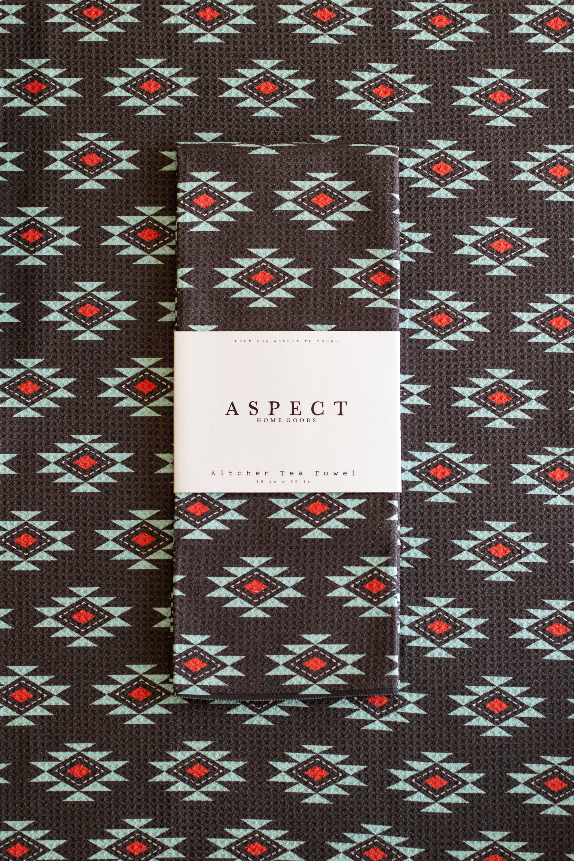 A folded waffle-weave kitchen tea towel with a geometric southwestern-style pattern, featuring a white paper belly band label in the center that reads 'ASPECT HOME GOODS' and 'KITCHEN TEA TOWEL'. The towel has a black background with repeating diamond motifs in turquoise, white, and red.