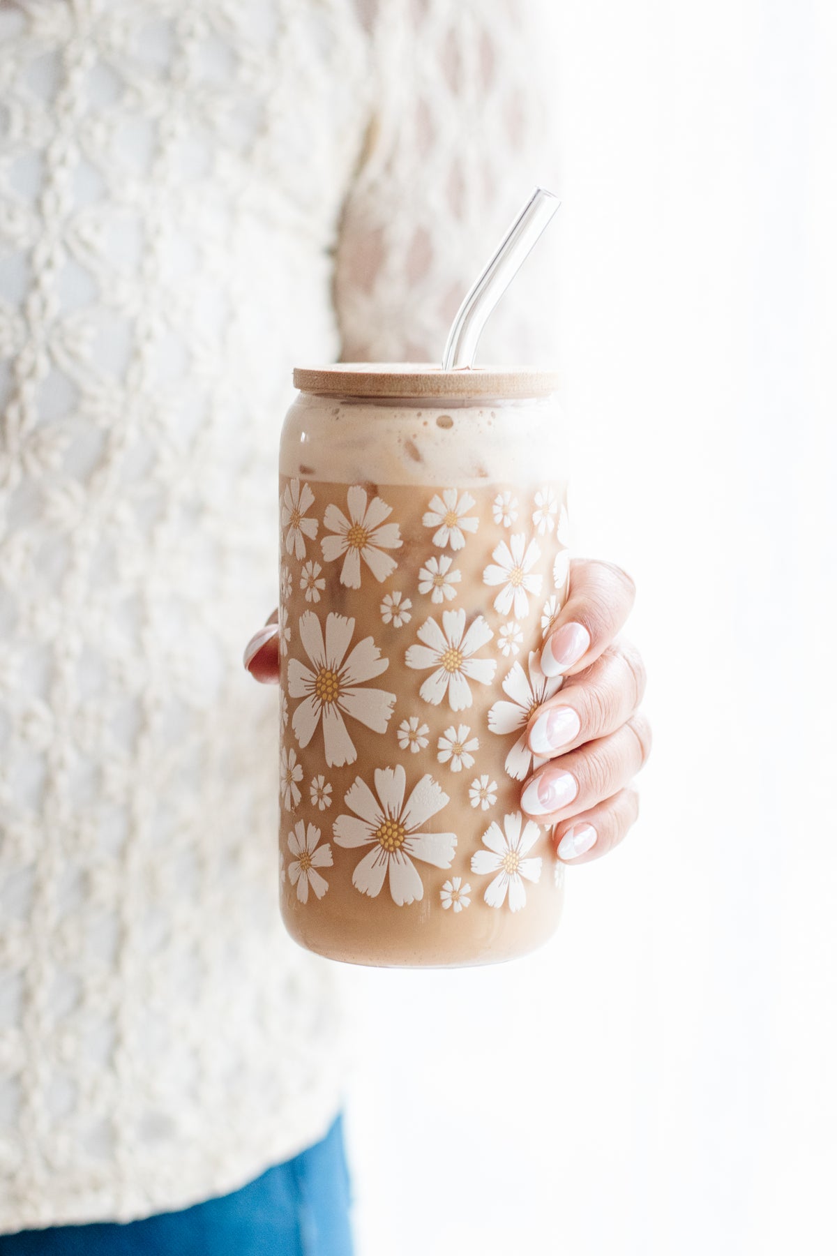 Person holding a white daisy floral-patterned can glass with a glass straw and a wooden bamboo lid, against a white background