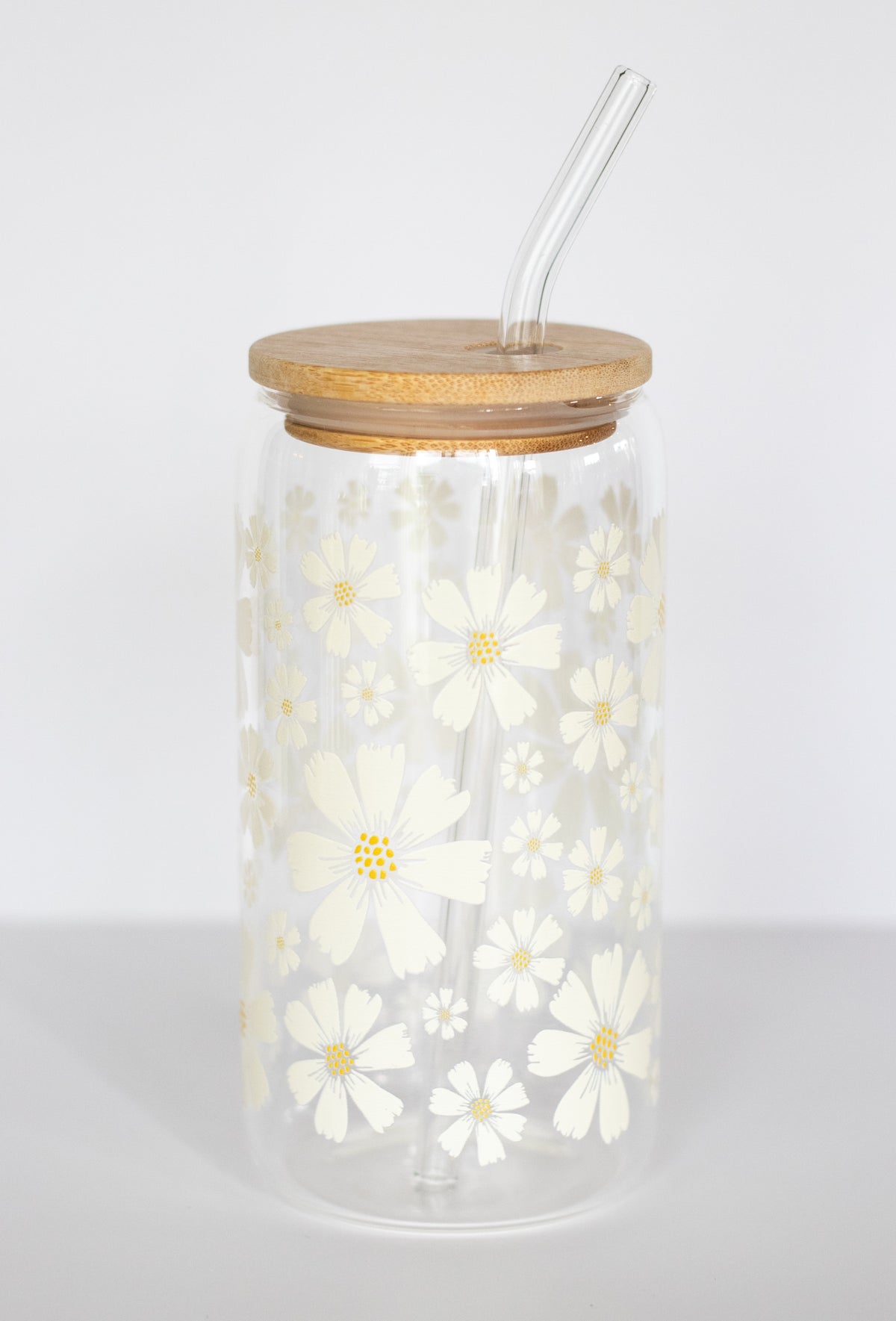 A can glass tumbler with a wooden bamboo lid and a glass straw, featuring a white daisy floral design against a white background.