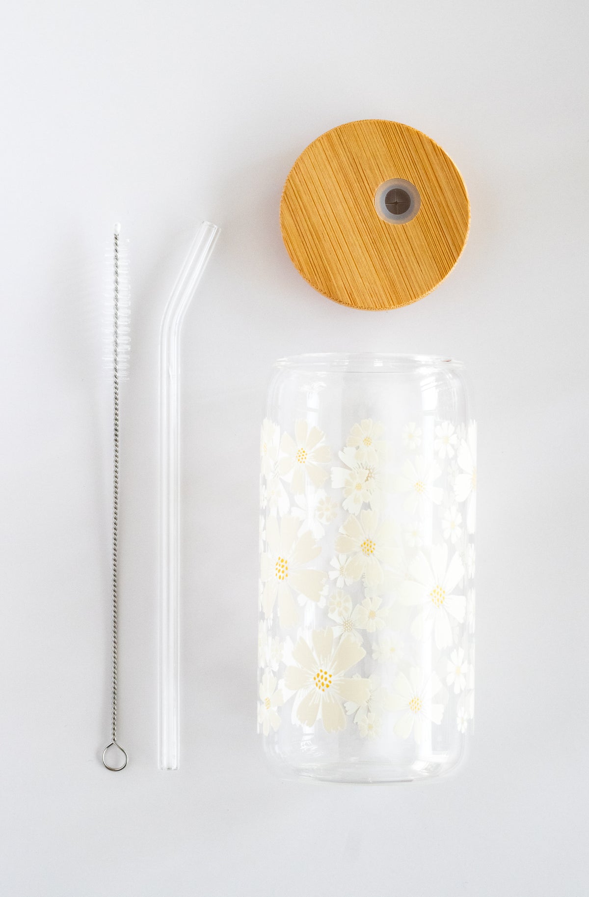 Clear can glass with a white daisy floral design, bamboo lid, a glass straw, and a straw brush cleaner, on a white background
