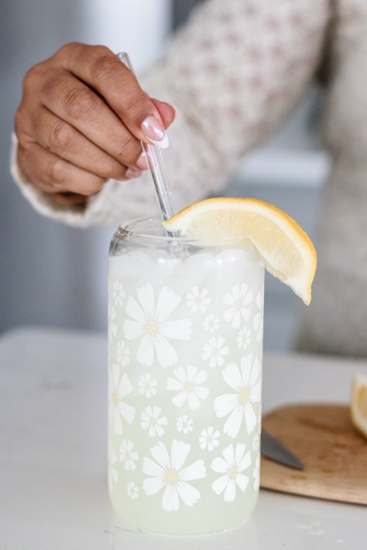 Hand using a glass straw to stir a beverage in a can glass with a white daisy floral design and a lemon wedge on a white surface.