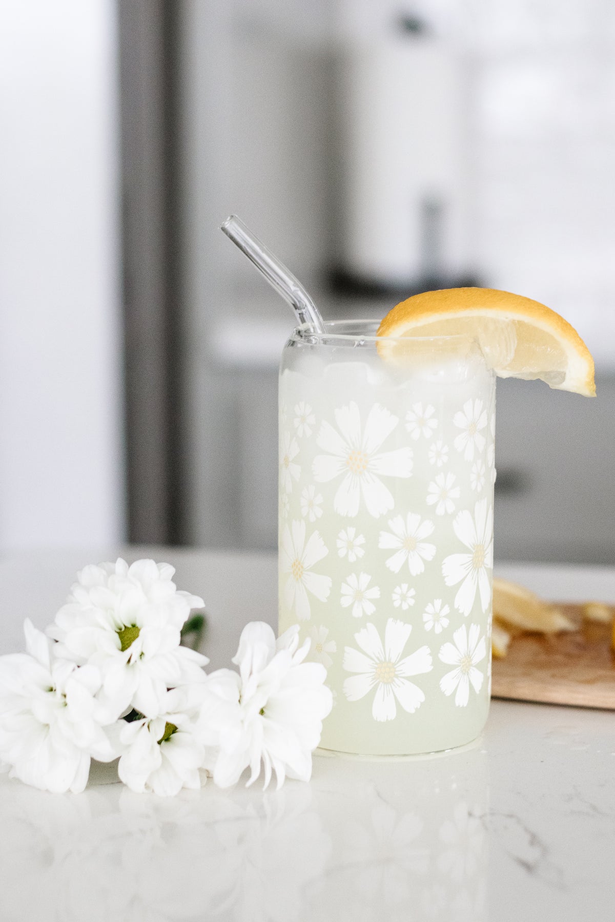 Can glass with a white daisy floral design, a lemon wedge, and a glass straw on a marble surface with white flowers.