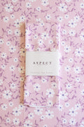 A folded kitchen tea towel with a pink, white, and purple floral pattern, wrapped in a white paper belly band labeled 'ASPECT HOME GOODS' and 'kitchen tea towel', laid flat on a matching patterned background.