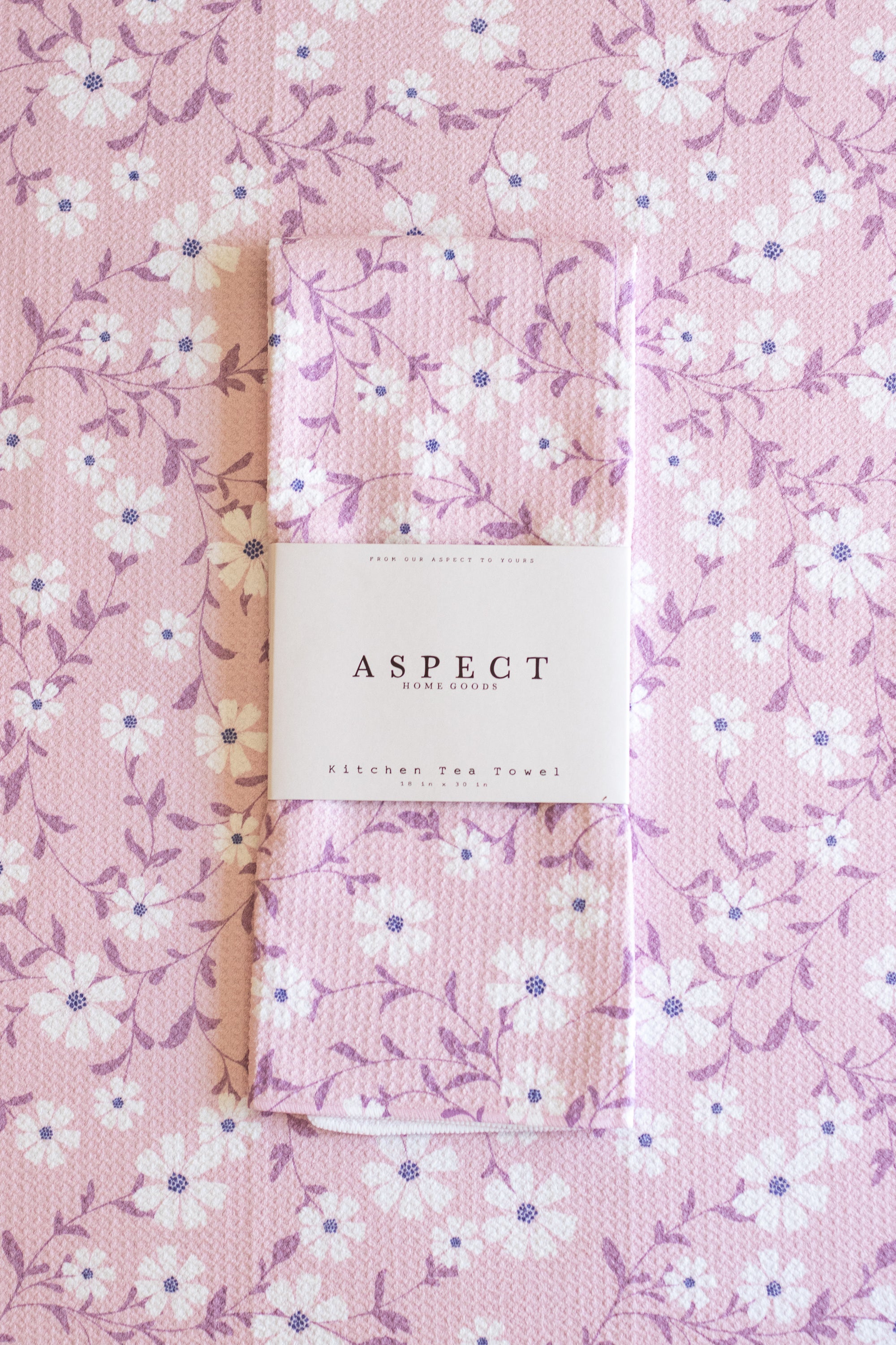 A folded kitchen tea towel with a pink, white, and purple floral pattern, wrapped in a white paper belly band labeled 'ASPECT HOME GOODS' and 'kitchen tea towel', laid flat on a matching patterned background.