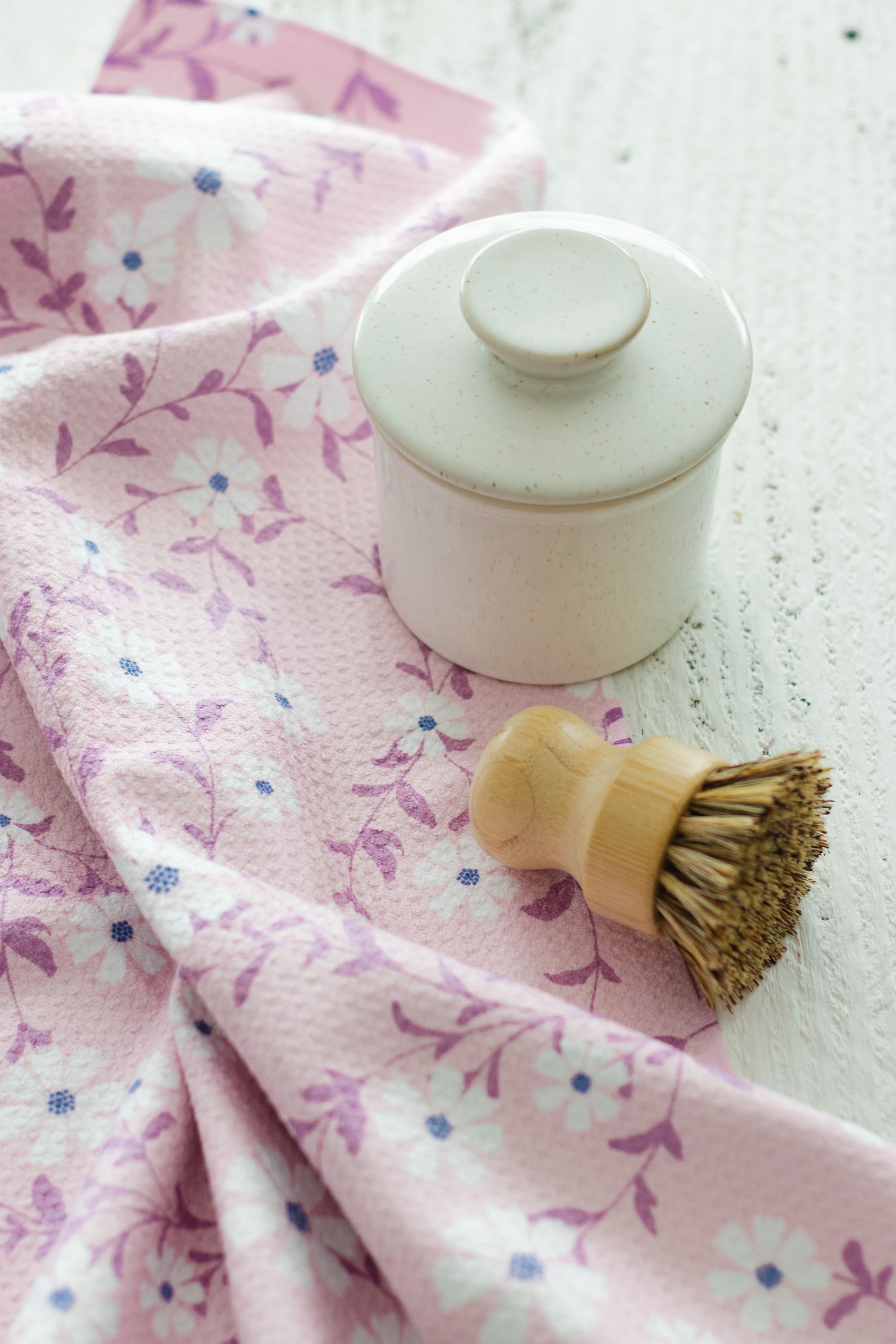 White ceramic butterbell jar with a wooden scrub brush on a pink floral waffle-weave kitchen tea towel against a textured white wooden surface.