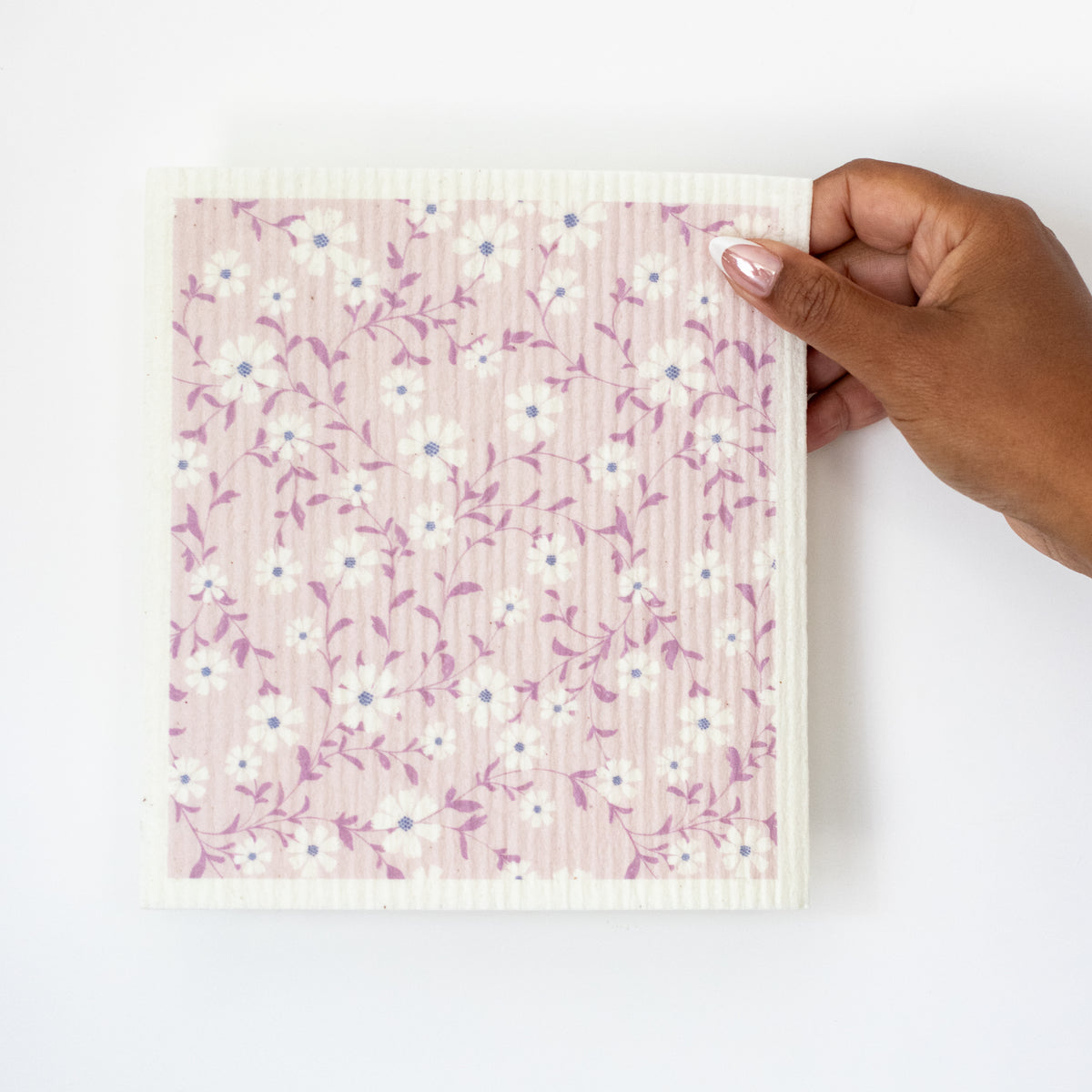 A hand holding a Swedish dishcloth with a white daisy pattern with purple leaves on a light pink background. 