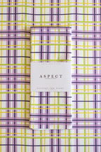 A folded waffle-weave kitchen tea towel with a purple, yellow, lime green, pink and white plaid pattern, wrapped in a white paper belly band labeled 'ASPECT HOME GOODS' and laid flat on a matching plaid background. 