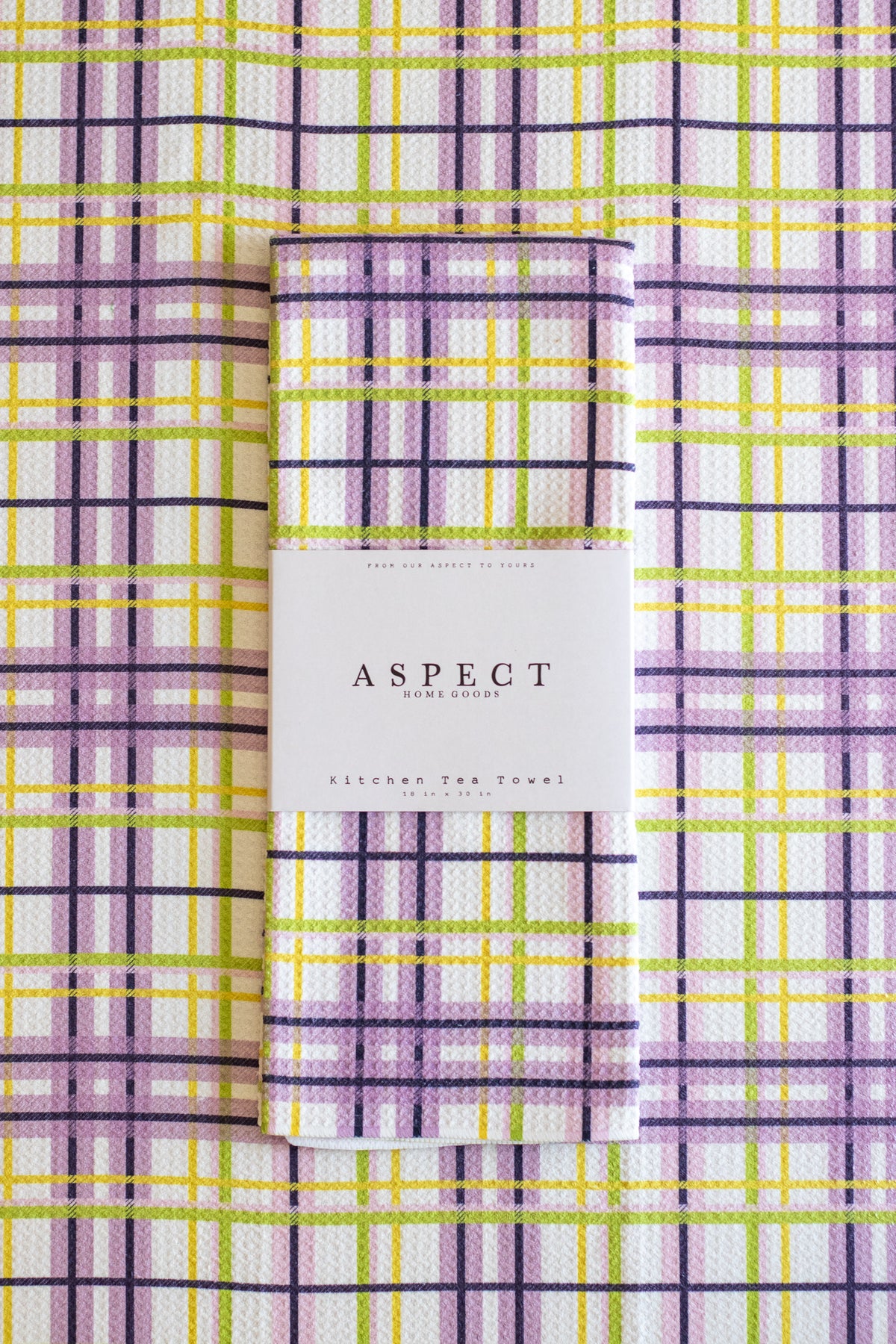 A folded waffle-weave kitchen tea towel with a purple, yellow, lime green, pink and white plaid pattern, wrapped in a white paper belly band labeled 'ASPECT HOME GOODS' and laid flat on a matching plaid background. 