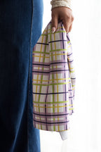 Person holding a plaid waffle-weave kitchen tea towel in a purple, green, yellow, and white color palette, against a white background.