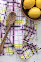 Wooden spoon on a plaid waffle-weave kitchen tea towel with lemons in a bowl, on a white wooden surface.