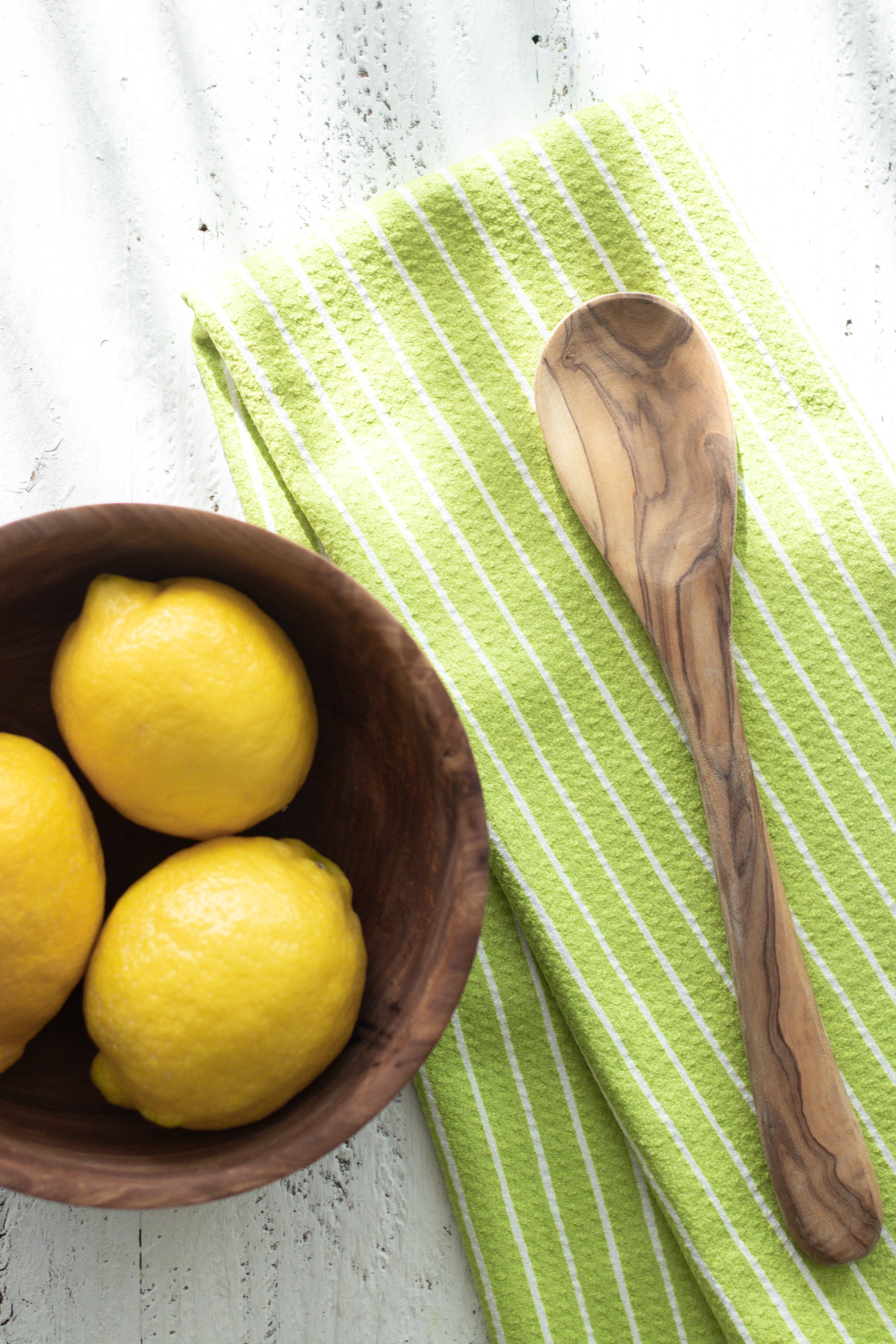 Bowl of lemons with a wooden spoon on a green striped waffle-weave kitchen tea towel against a textured white wooden surface.