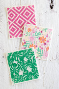 Three Swedish dishcloths with different prints are arranged on a white distressed wooden surface. One napkin features a pink-and-white geometric diamond pattern; another has a colorful floral print with orange, pink, and purple flowers and greenery; and the third displays a green monstera leaf pattern.