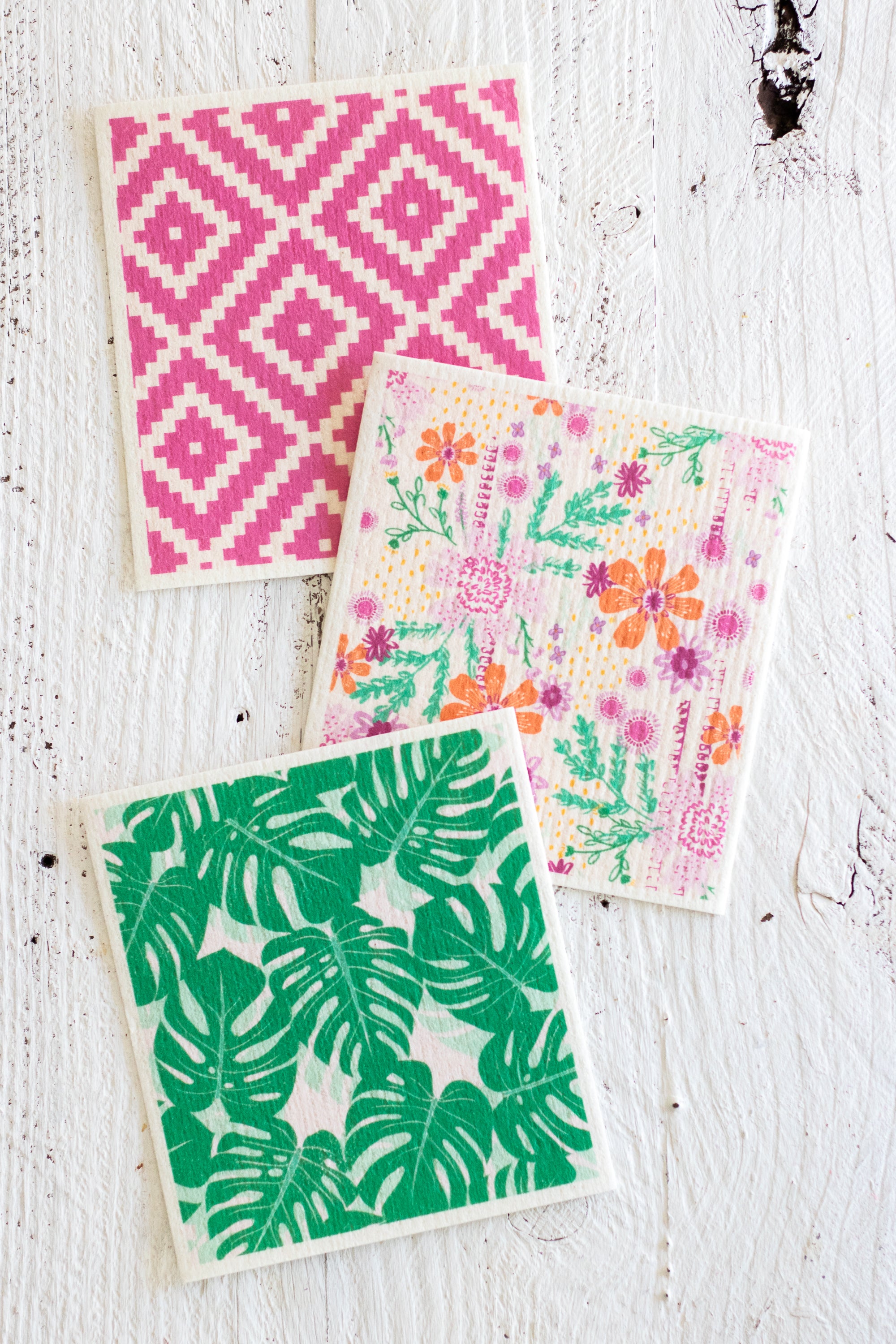 Three Swedish dishcloths with different prints are arranged on a white distressed wooden surface. One napkin features a pink-and-white geometric diamond pattern; another has a colorful floral print with orange, pink, and purple flowers and greenery; and the third displays a green monstera leaf pattern.