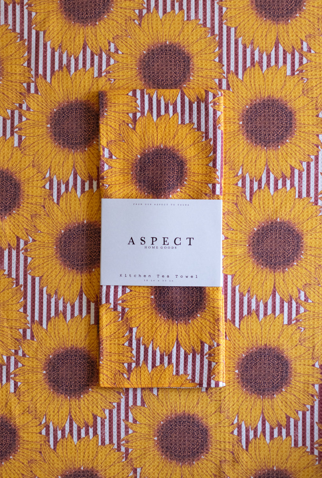 A folded waffle-weave kitchen tea towel with a yellow sunflower and burgundy stripe pattern, laid flat on top of a matching patterned background. A white paper belly band label in the center reads 'ASPECT Home Goods' and 'Kitchen Tea Towel'.