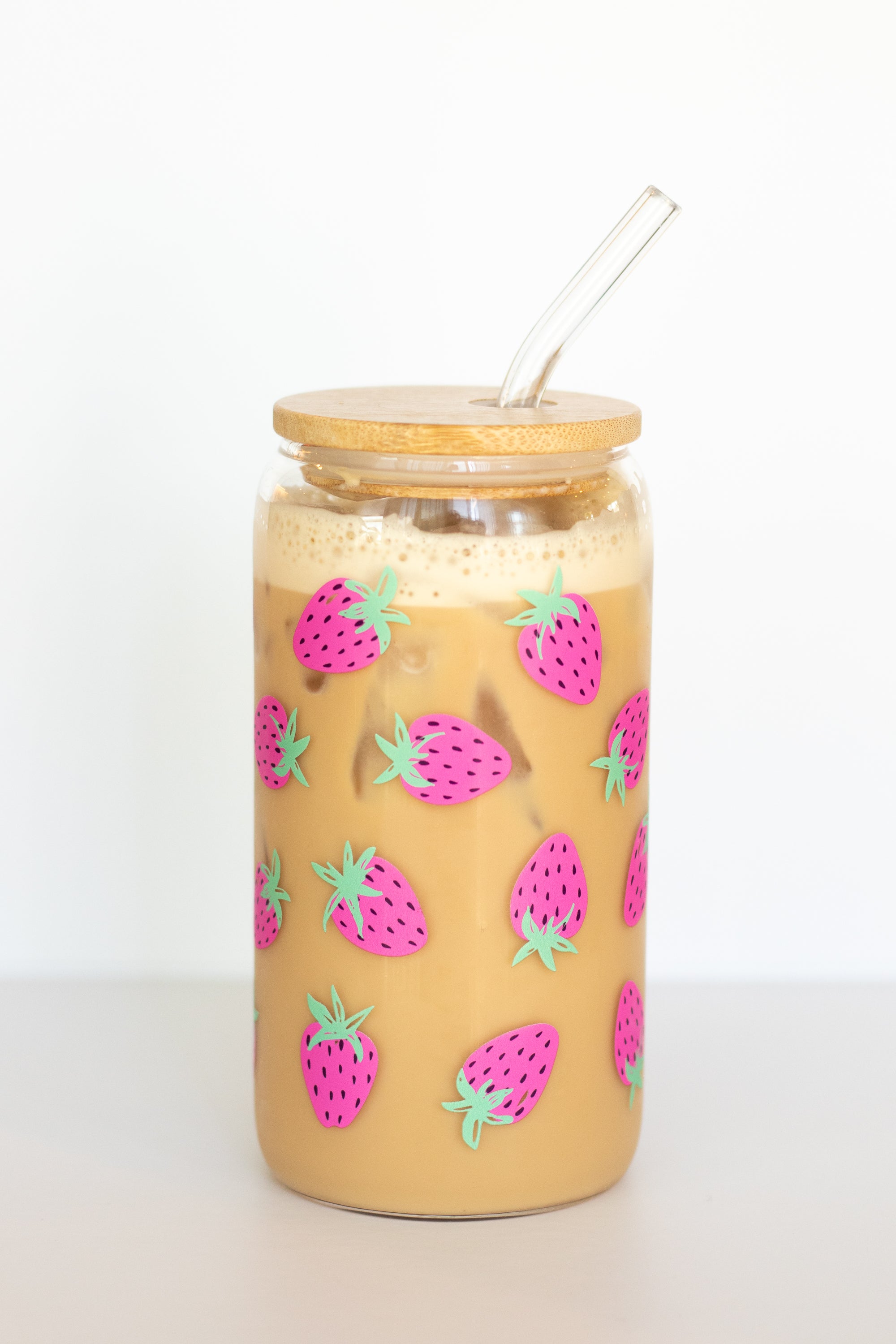A can glass tumbler that is decorated with hand-painted pink-and-green strawberries, with a wooden lid and a glass straw, containing an iced coffee drink, against a white background.