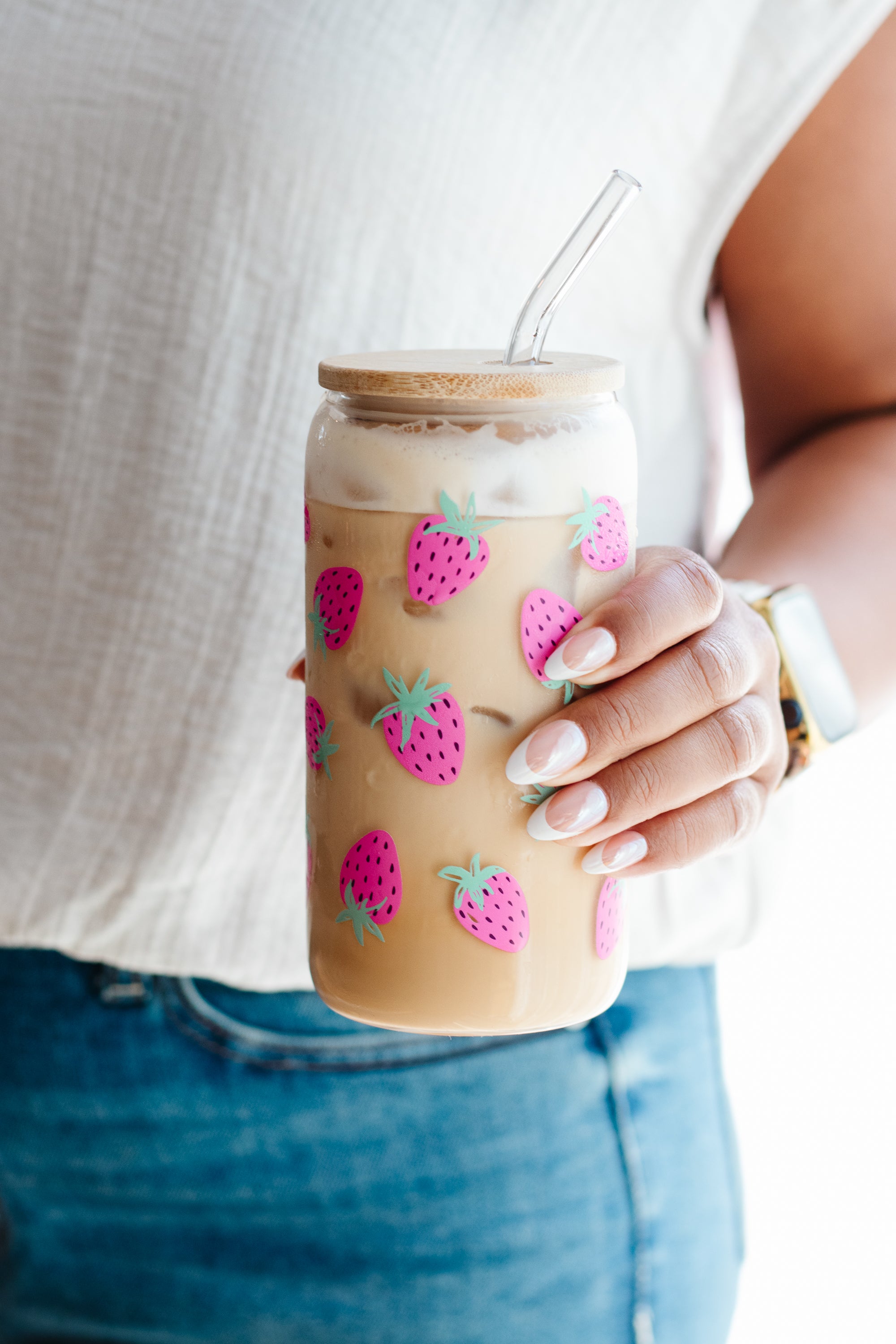 A person holding a clear glass can-style tumbler with a bamboo lid and clear straw, filled with an iced coffee drink. The tumbler is decorated with pink-and-green strawberry illustrations.