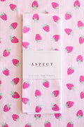 A folded kitchen tea towel with a pink strawberry pattern, wrapped in a white paper belly band labeled 'ASPECT HOME GOODS' and 'Kitchen Tea Towel', set against a matching flat-lay background of the same towel.