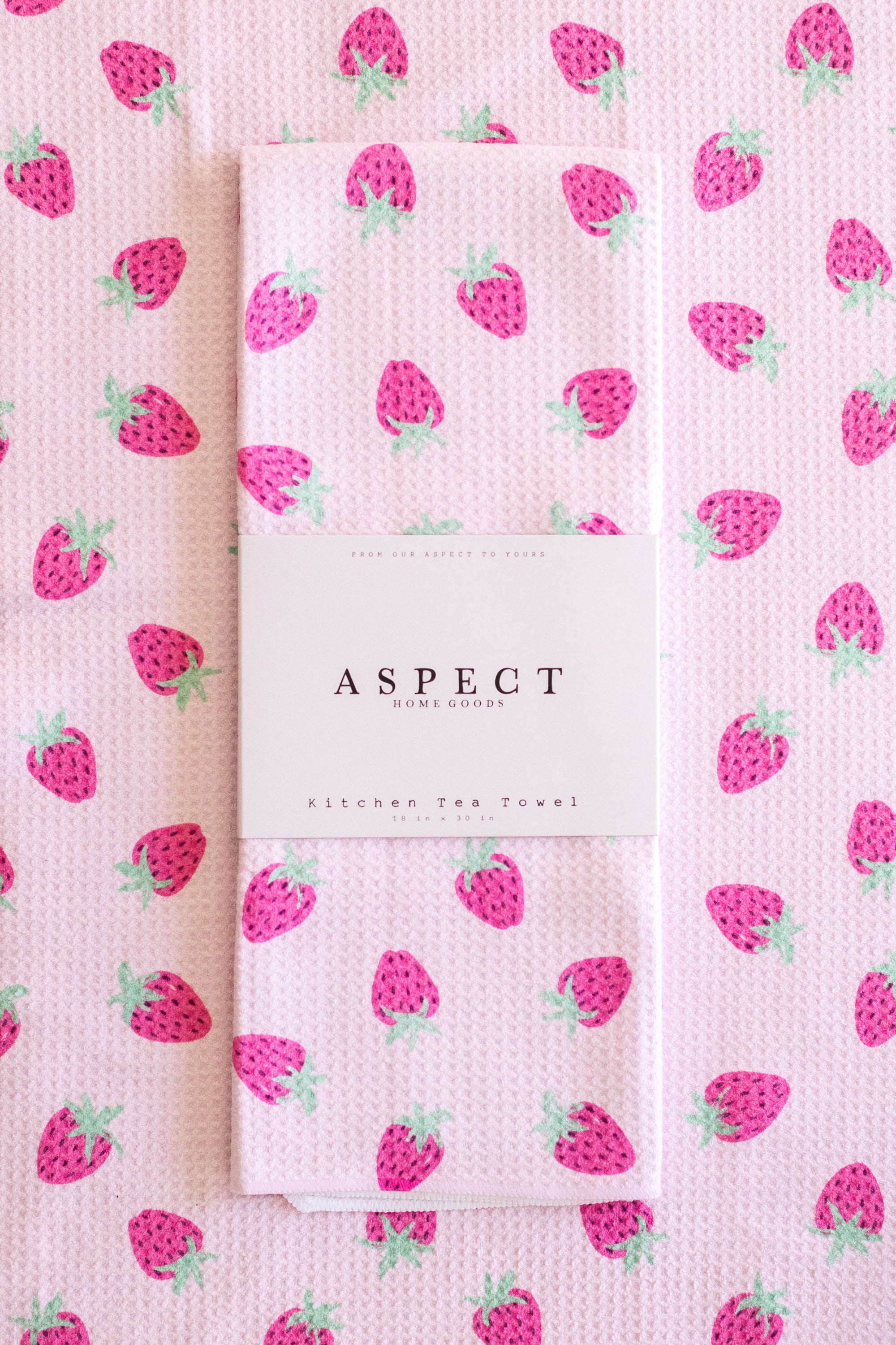 A folded kitchen tea towel with a pink strawberry pattern, wrapped in a white paper belly band labeled 'ASPECT HOME GOODS' and 'Kitchen Tea Towel', set against a matching flat-lay background of the same towel.