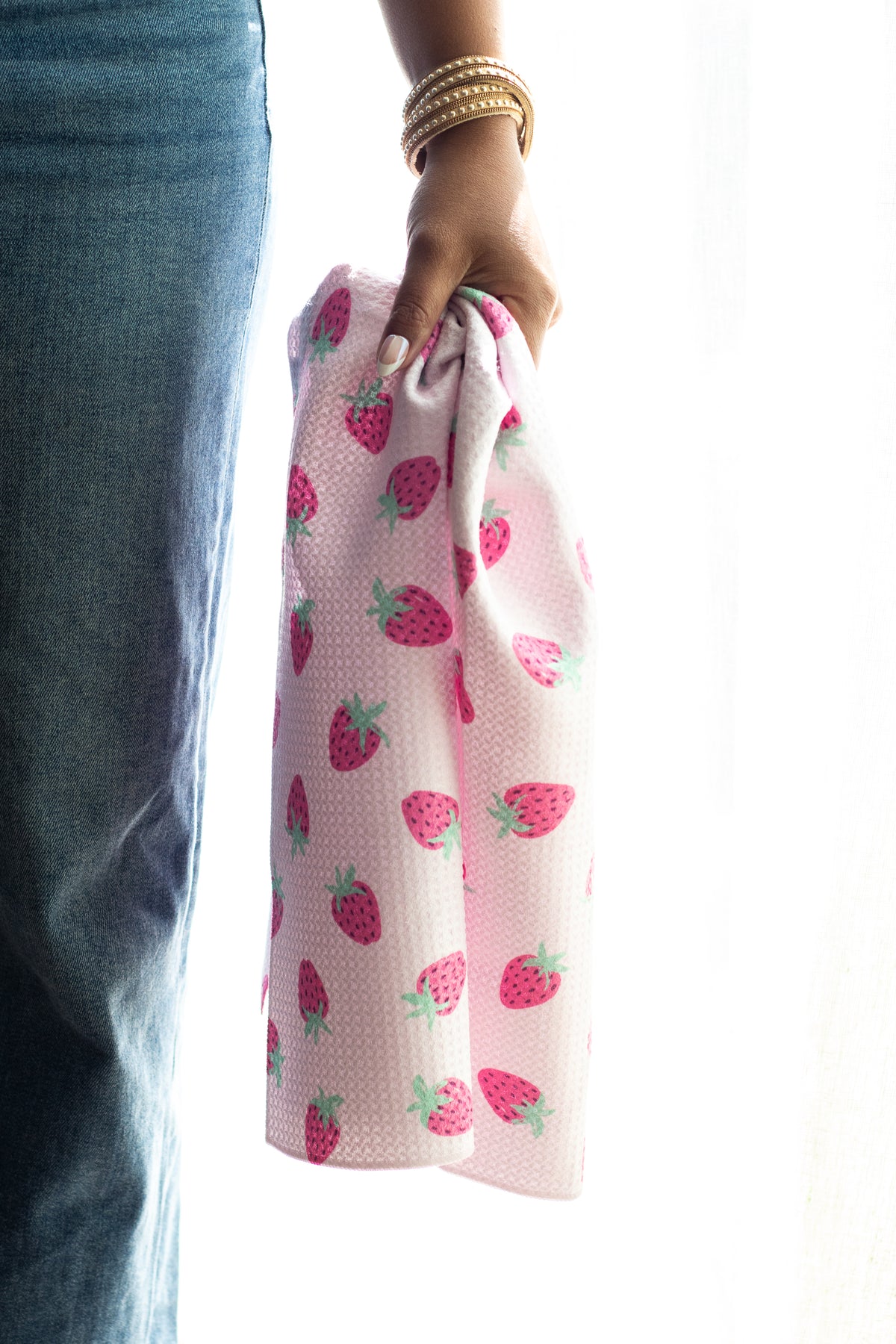 A person holding a light pink waffle-weave kitchen tea towel with a repeating strawberry pattern, next to blue jeans against a bright white background.