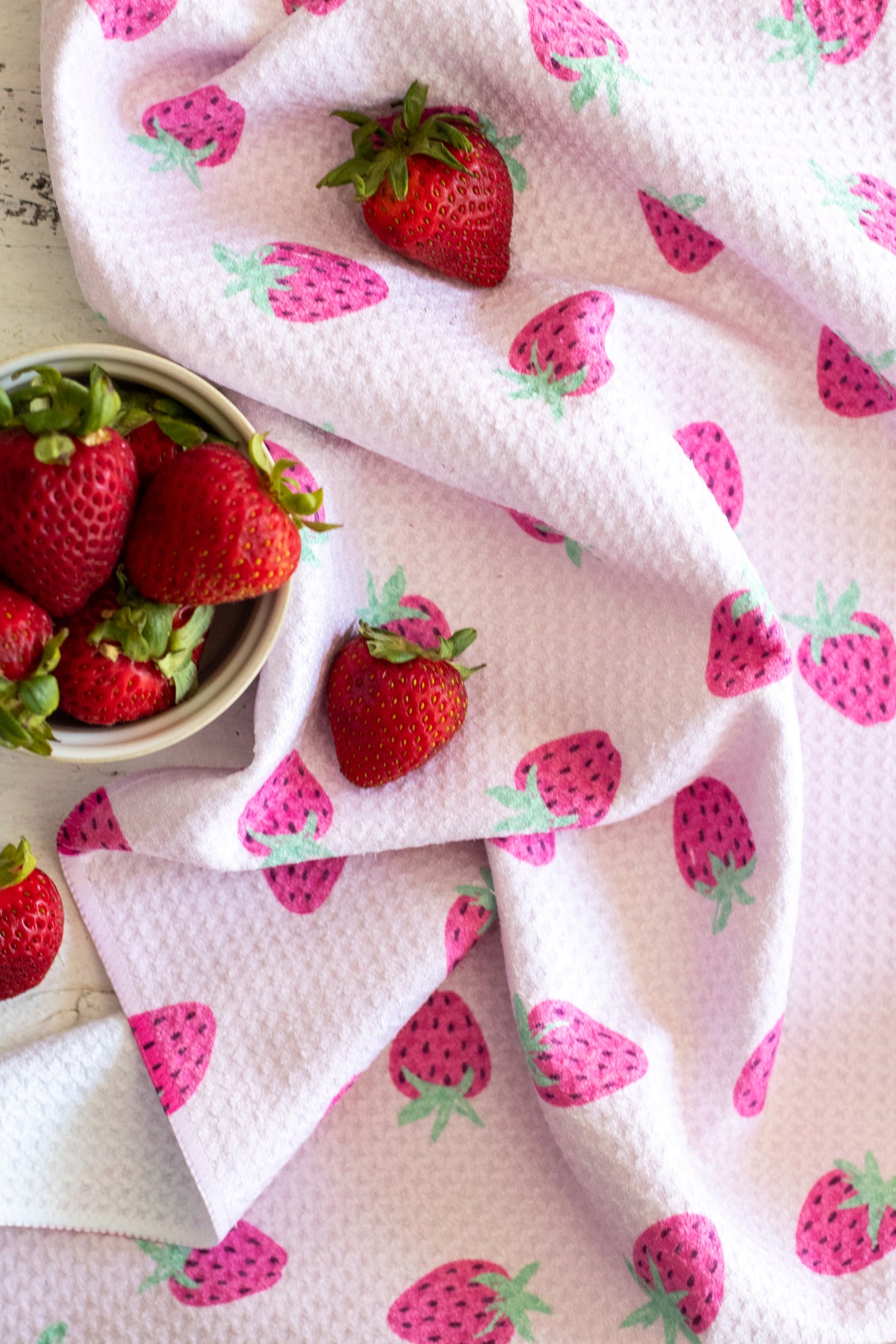 A white waffle-weave kitchen towel with a pink-and-green strawberry pattern, styled with fresh strawberries in a small bowl and scattered on a white wooden surface.