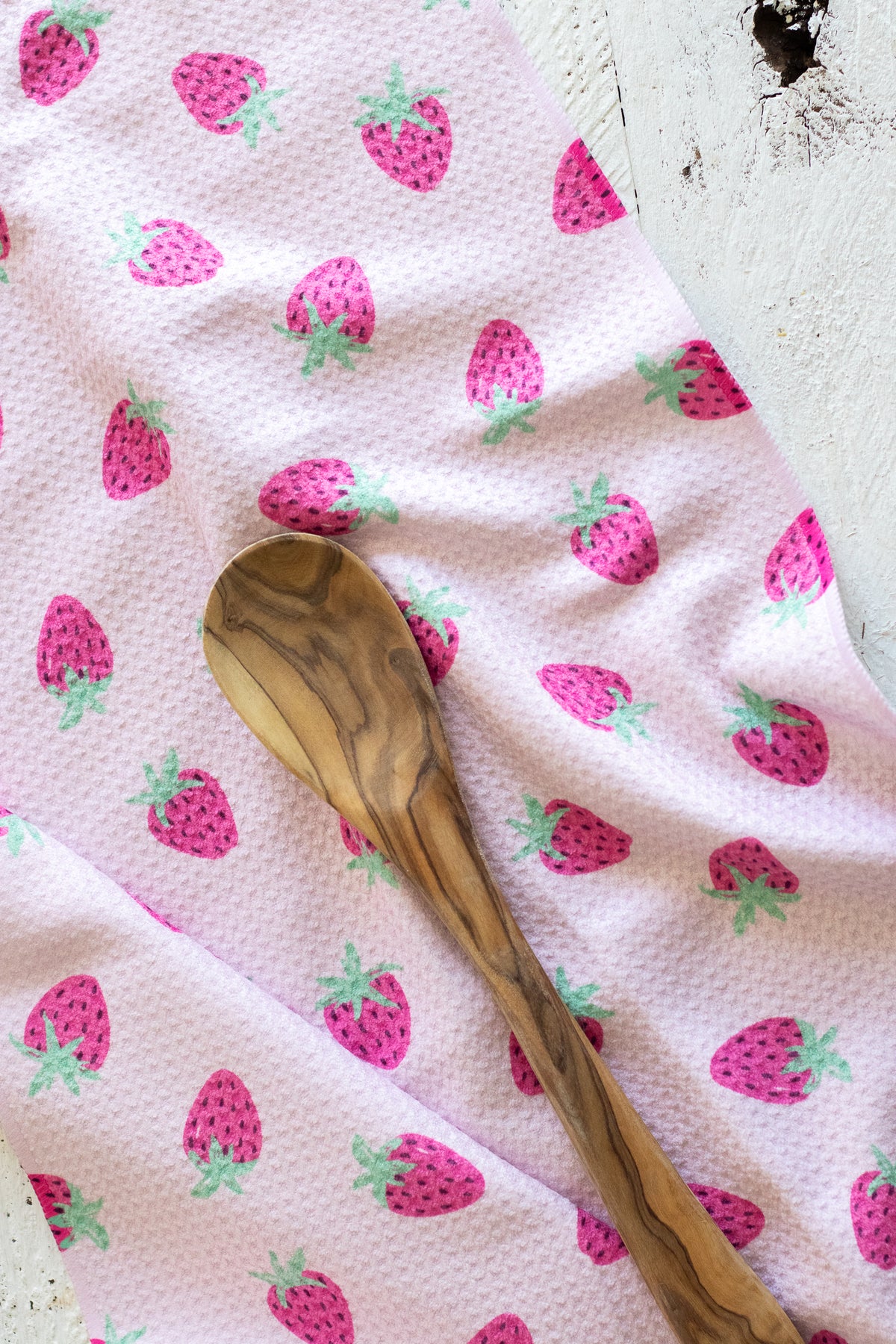 A close-up of a light pink waffle-weave kitchen tea towel with a repeating pattern of pink strawberries with green leaves, styled with a wooden spoon on a rustic white wooden surface.