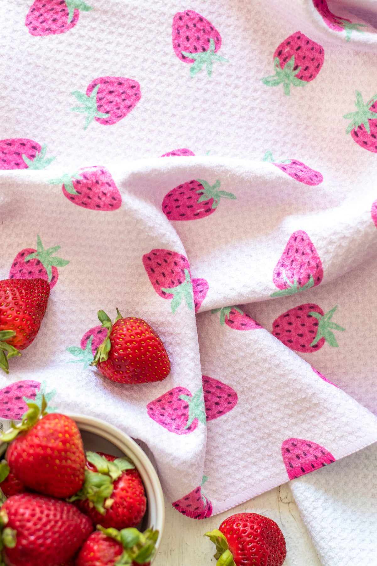 A light pink waffle-weave kitchen towel with a pink-and-green strawberry pattern, styled with fresh red strawberries on a white wooden surface.