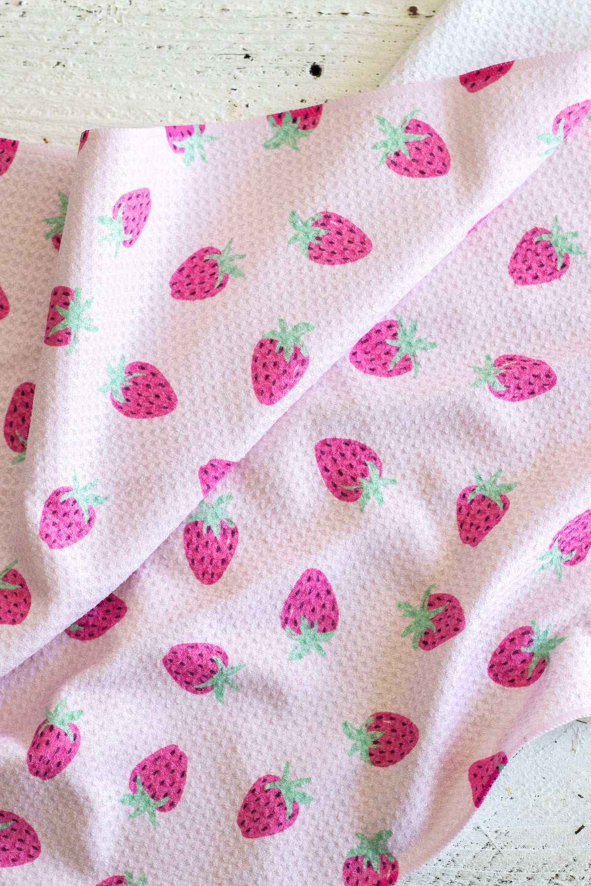 A close-up of a light pink waffle-weave kitchen tea towel with a repeating pattern of pink strawberries with green leafy tops, draped over a white rustic wooden surface.