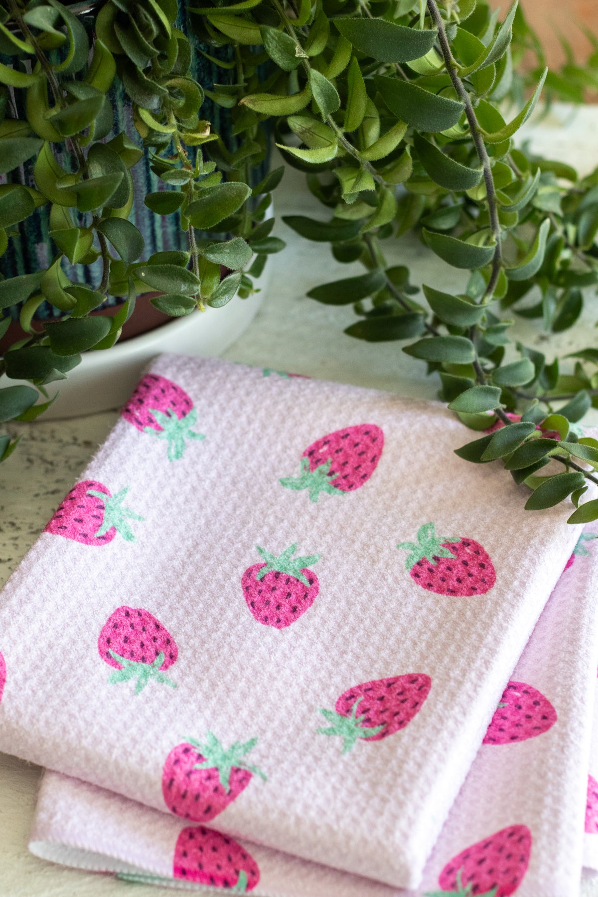 A folded light pink waffle-weave kitchen towel with a pink-and-green strawberry pattern, styled with green trailing houseplants in the background.